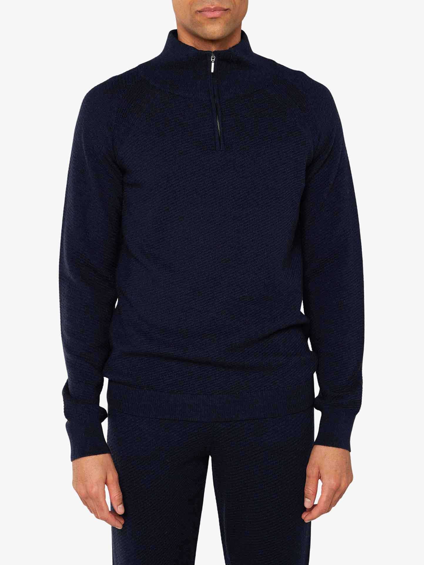 The person is wearing the We Norwegians Geilo 1/4-Zip Men, an elegant navy blue long-sleeved sweater crafted from fine Merino wool. The sweater boasts a textured pattern and a stand-up collar with a zipper that extends halfway down the chest. Paired with matching navy blue pants, this chic lounge set stands out against the plain white background.