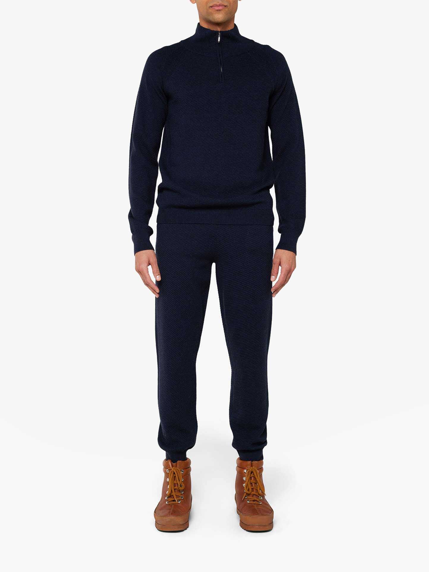 A person is wearing an elegant Geilo 1/4-Zip Men, a dark navy blue long-sleeve pullover from We Norwegians, paired with matching navy blue jogger pants. They are also sporting brown lace-up boots. The chic lounge set stands out against the plain white background.