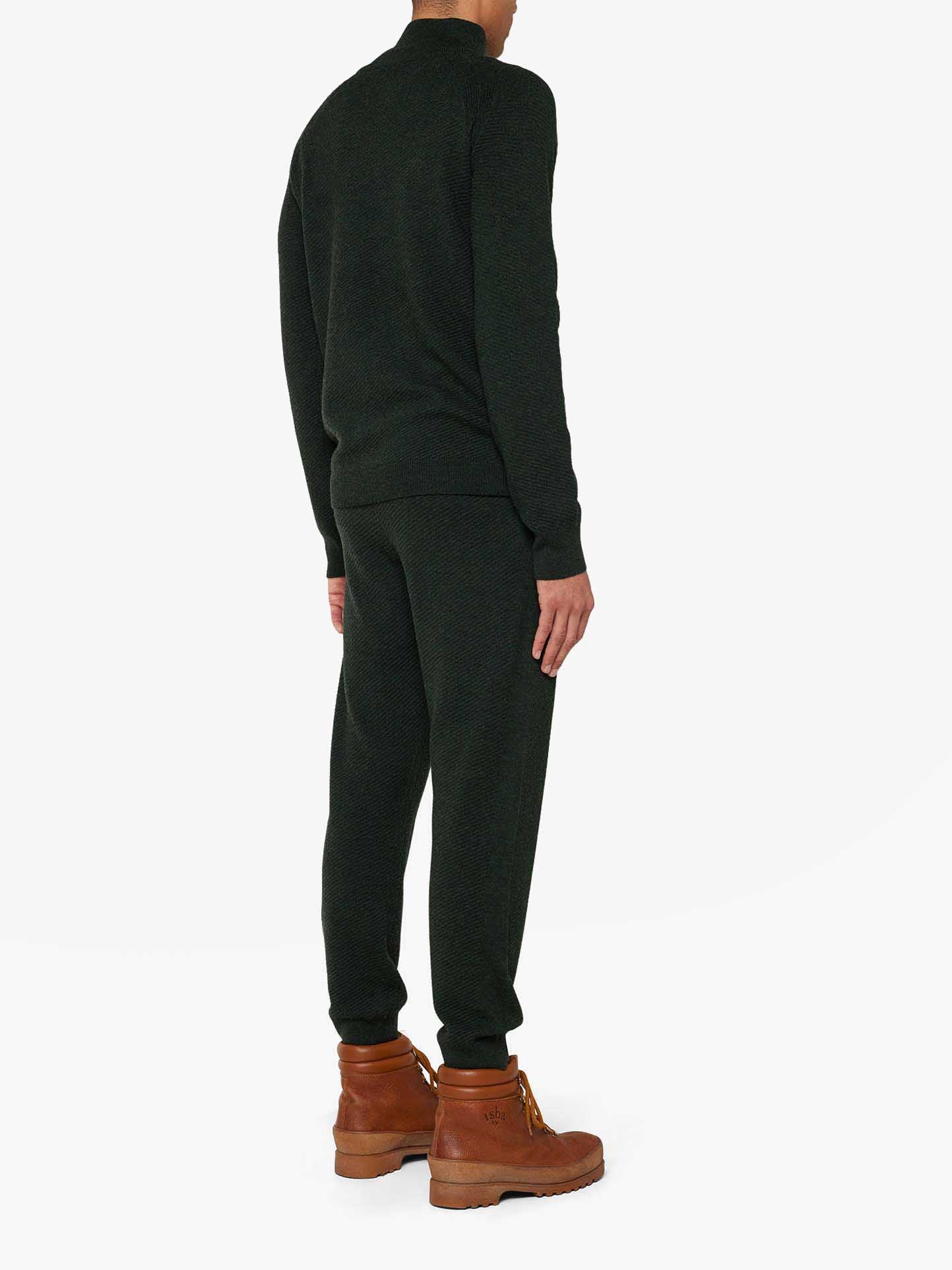 A person stands with their back to the camera, dressed in a chic Geilo 1/4-Zip Men lounge set from We Norwegians and brown leather boots. The dark green sweatsuit contrasts against the white background, making them and their stylish attire the focal point of the image.