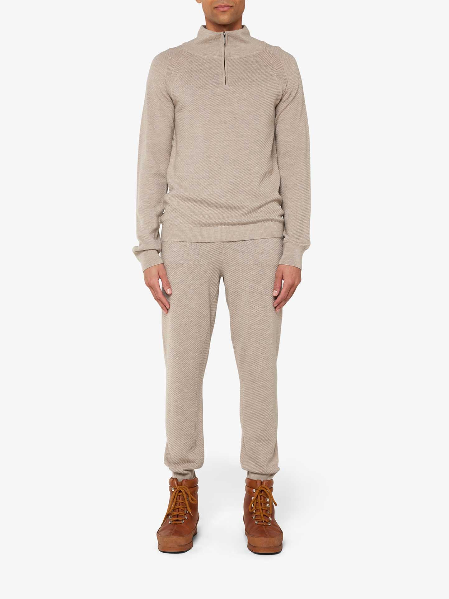 A person stands wearing the elegant Geilo 1/4-Zip Men by We Norwegians, a beige long-sleeve pullover paired with matching jogger-style pants, exuding comfort and sophistication. The outfit is complemented by brown lace-up boots against a plain white background.