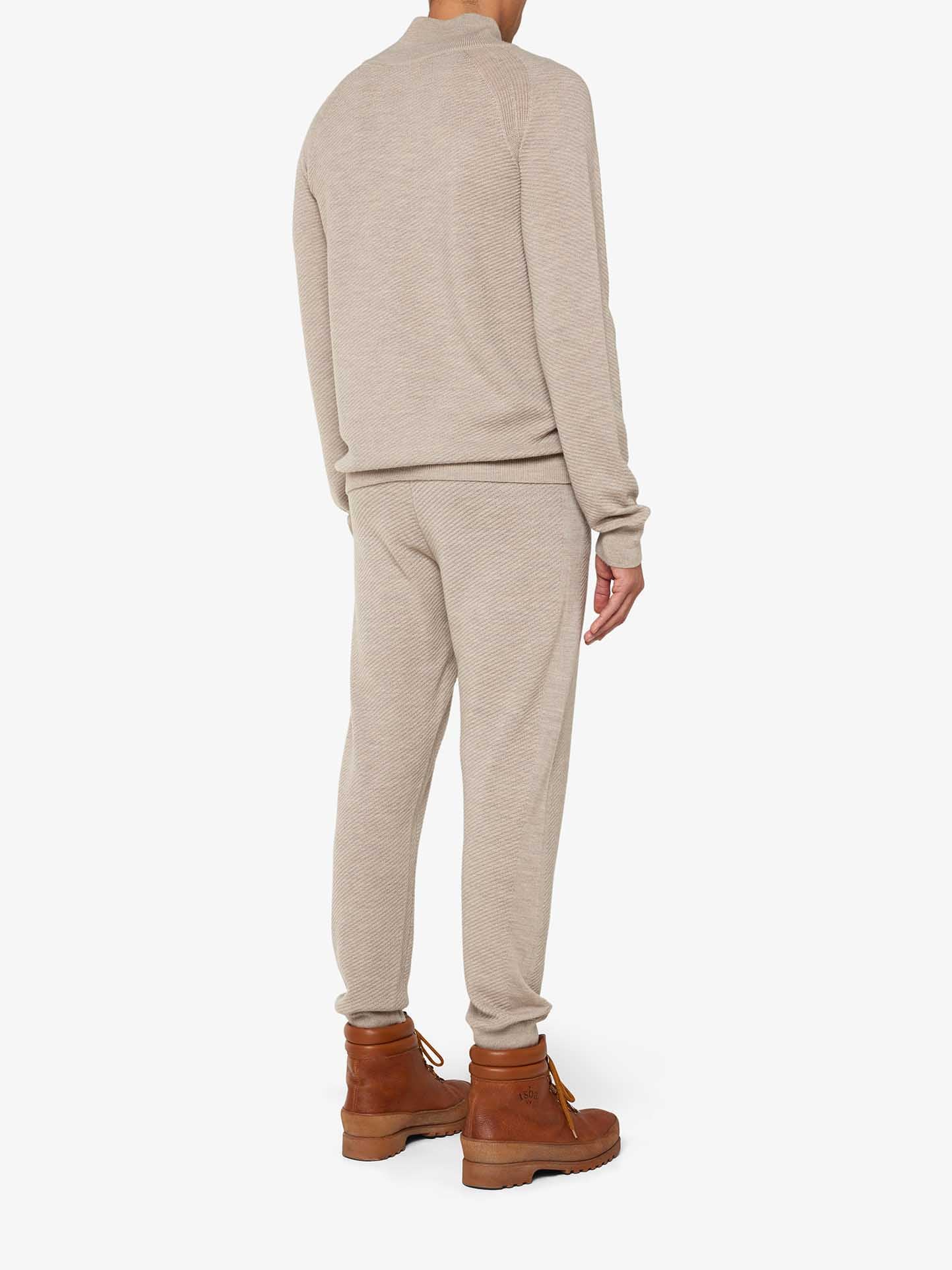 A person stands facing away, showcasing a chic lounge set featuring the We Norwegians Geilo 1/4-Zip Men in beige, with matching long-sleeve top and pants made from Fine Merino Wool, complemented by brown boots. The background is plain white.