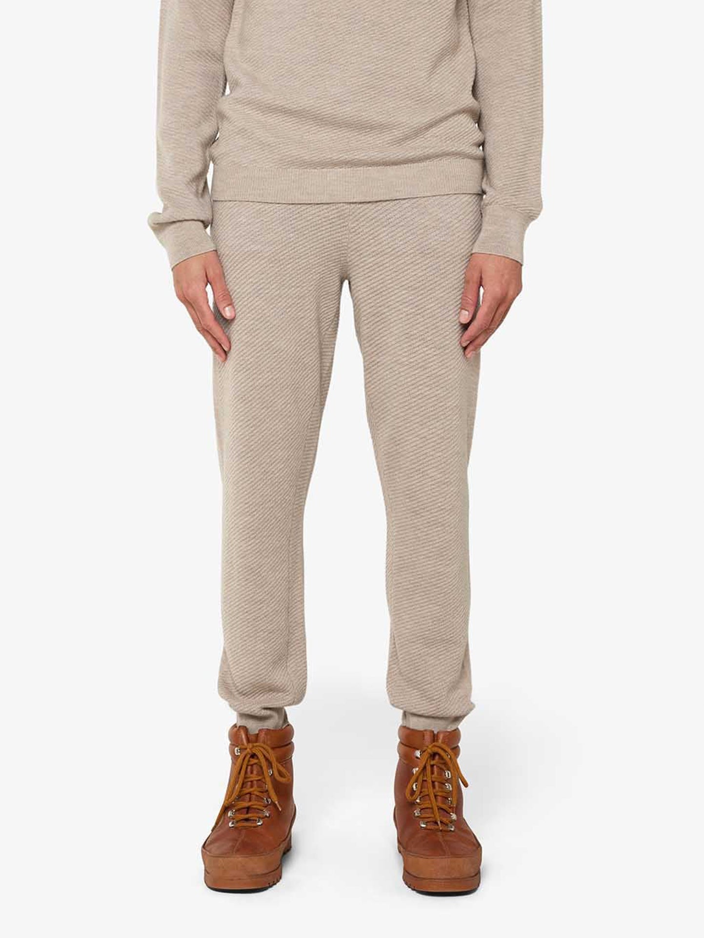 A person is shown from the shoulders down, wearing a light beige long-sleeved top and Geilo Jogger Men in matching Merino wool with ribbed cuffs by We Norwegians. They are also wearing brown ankle-high boots with laces. The background is plain white.