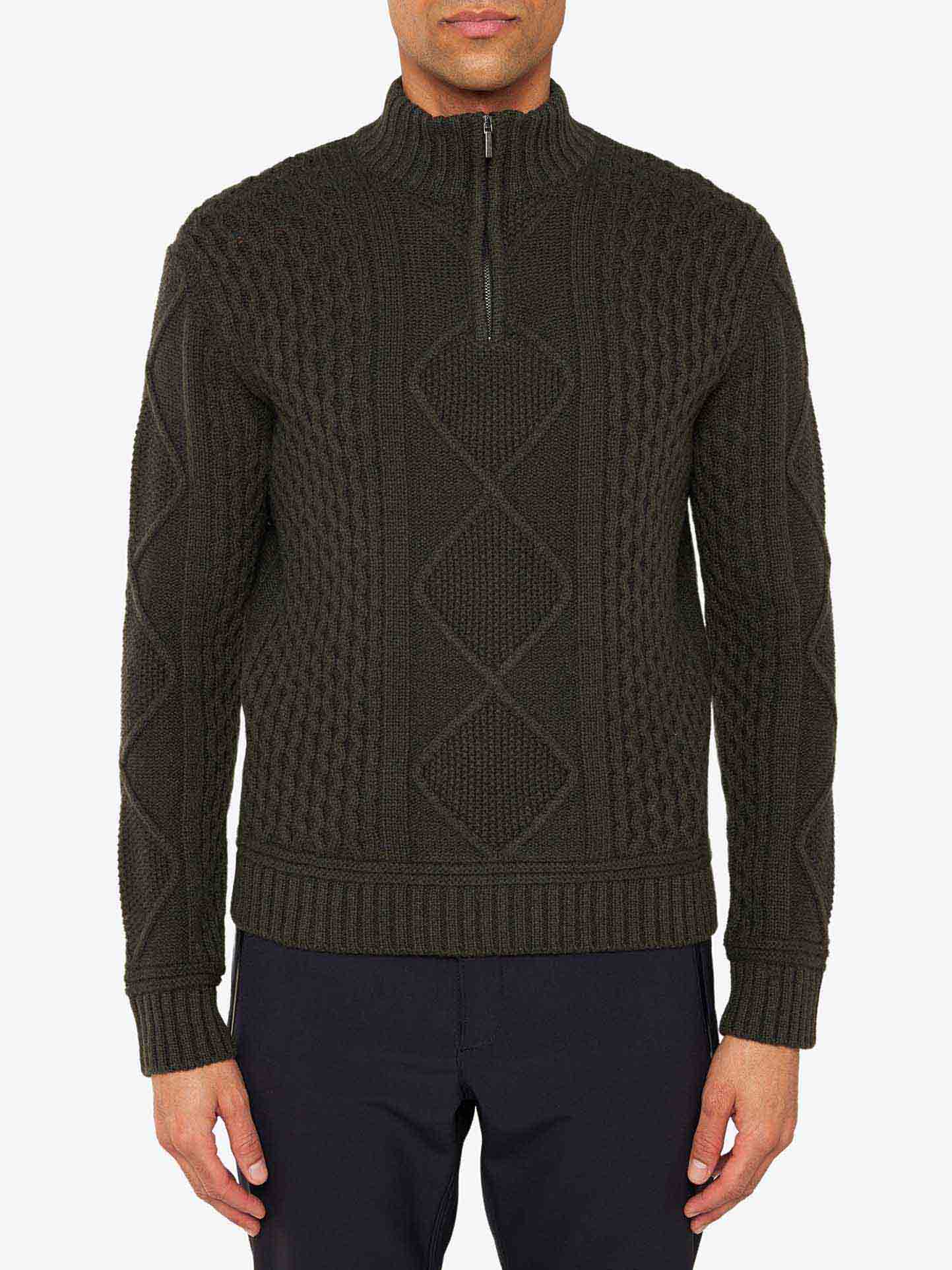 A person wearing the Geilo Cableknit 1/4-Zip Men sweater by We Norwegians, crafted from luxurious Italian yarn in a dark green color. The sweater showcases a large diamond pattern on the front and intricate cable knit designs on the sides and sleeves. Paired with dark pants, the high quarter zip collar adds a stylish touch to the ensemble.