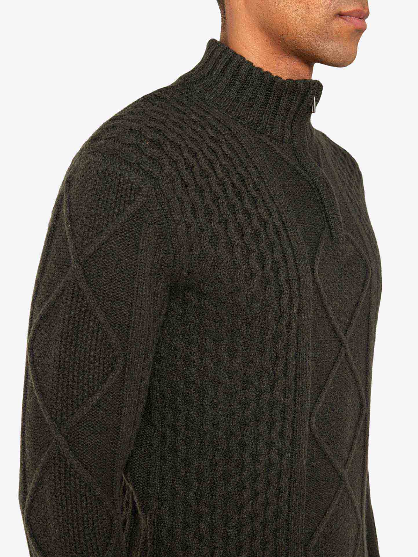 A person wearing the Geilo Cableknit 1/4-Zip Men by We Norwegians, a dark green sweater made from Italian yarn, featuring a high collar and zipper detail. The Merino wool garment showcases a blend of diamond and braided patterns. The person's face is not fully visible against the plain white background.