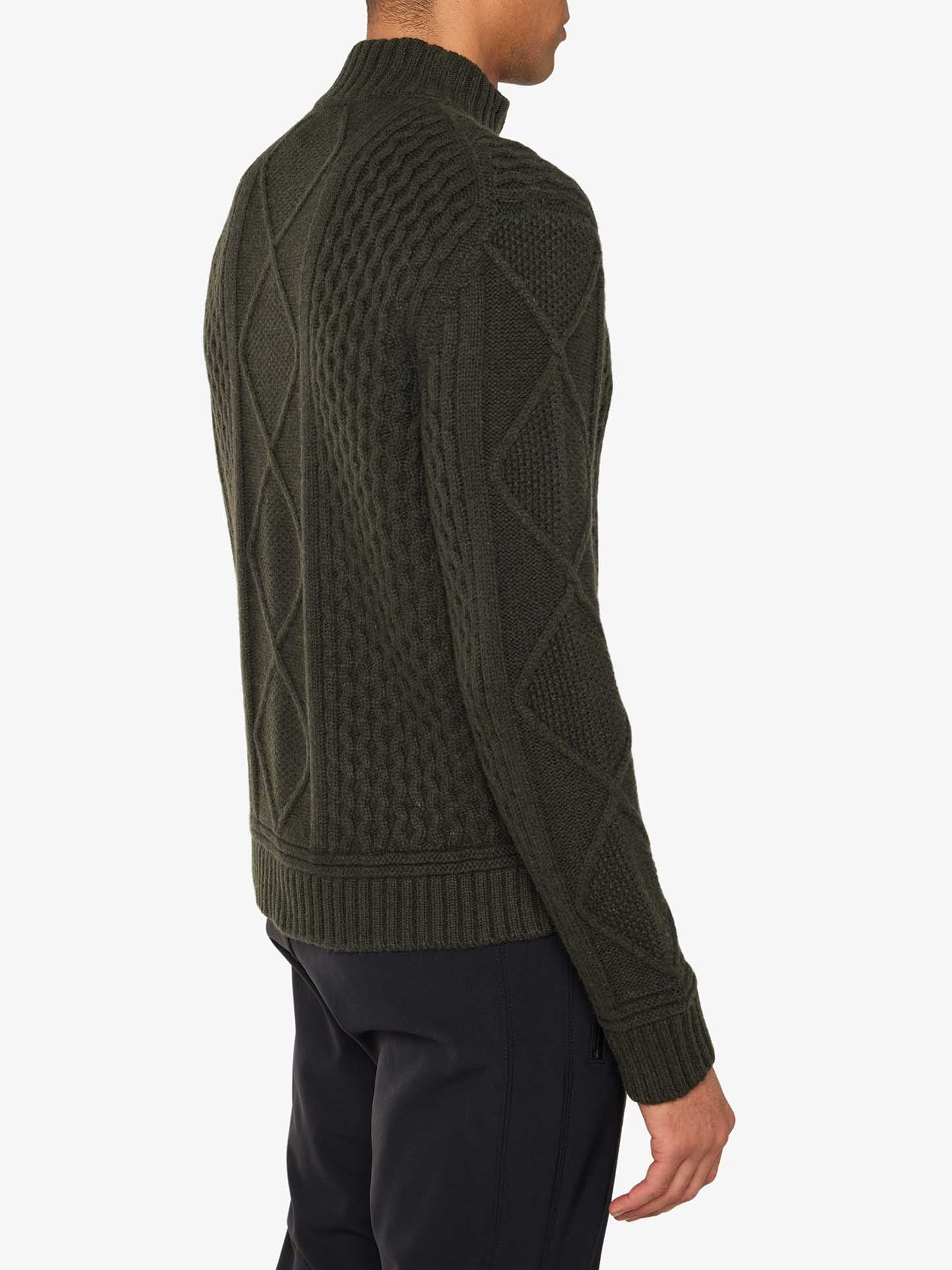 A person is wearing the Geilo Cableknit 1/4-Zip Men from We Norwegians, made of luxurious Italian yarn, featuring diamond and braided patterns in dark green. The high collar and ribbed cuffs and hem complete the look. They are standing sideways, facing to the right, paired with black pants for a timeless ensemble.