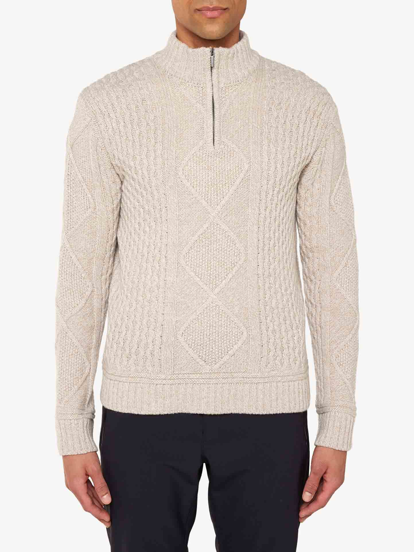 A person is wearing the Geilo Cableknit 1/4-Zip Men by We Norwegians, a beige, cable-knit sweater with a high collar and a half-zip front, crafted from fine Merino Wool. The sweater features intricate diamond and braid patterns. Paired with black pants, the elegant ensemble stands out against the plain white background.