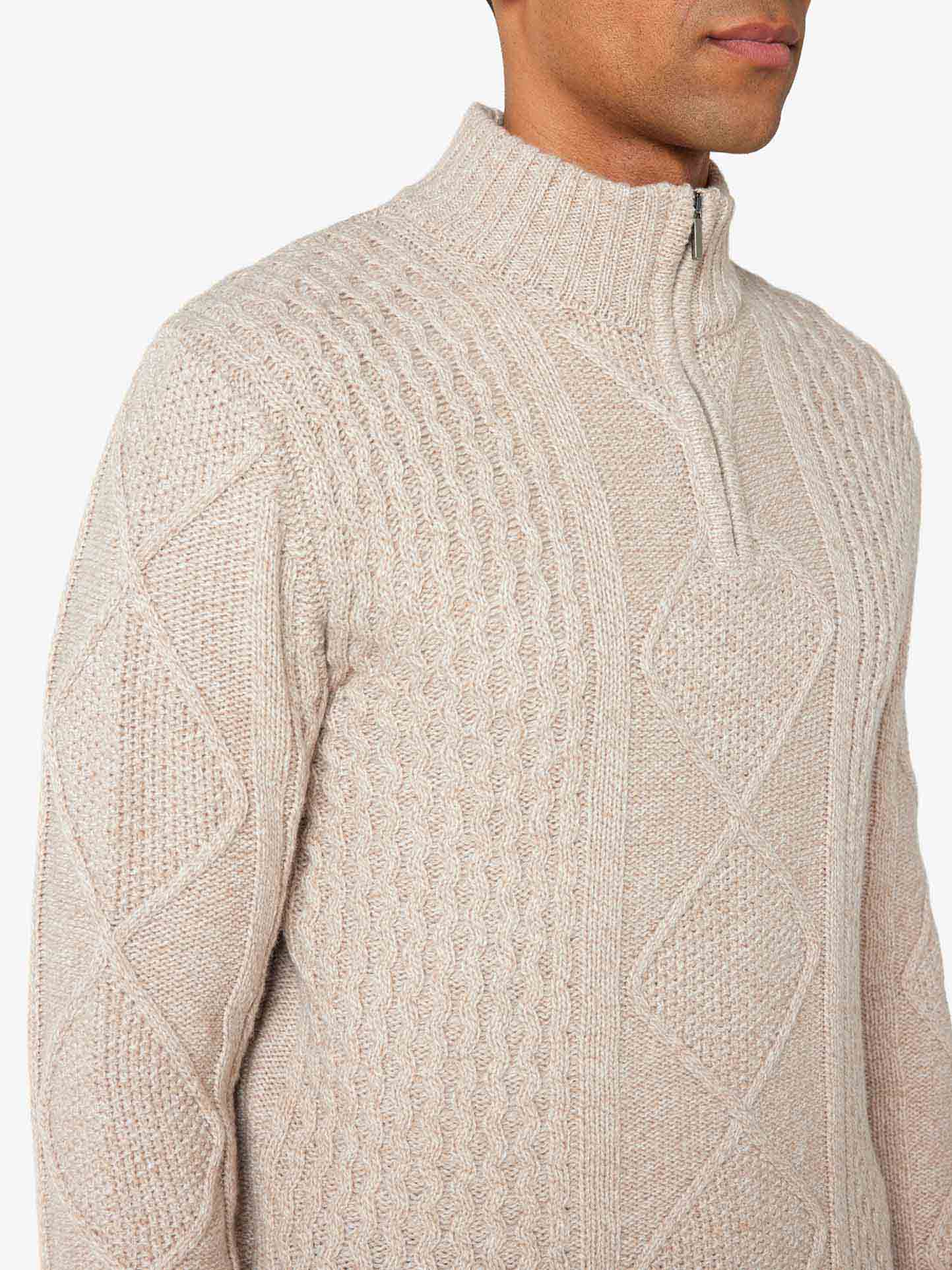 A person wearing the "Geilo Cableknit 1/4-Zip Men" by We Norwegians, a beige chunky knit sweater made of soft Merino Wool with a high collar and quarter-zip. The sweater features intricate patterns, including classic cable knits and diamond shapes, crafted from luxurious Italian yarn. The image shows the upper half of the person against a plain background.