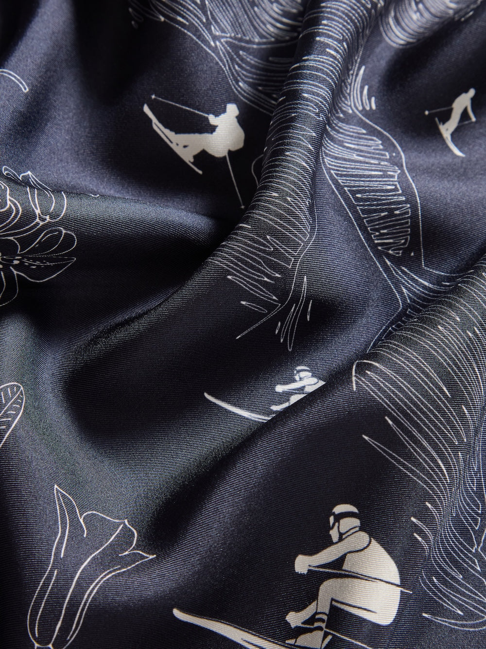 Close-up of the We Norwegians Alps Silk Scarf, featuring Scandinavian-inspired white line drawings of skiing, snowboarding, and wakeboarding among abstract shapes and botanical details. Soft folds add elegant shadows and texture.