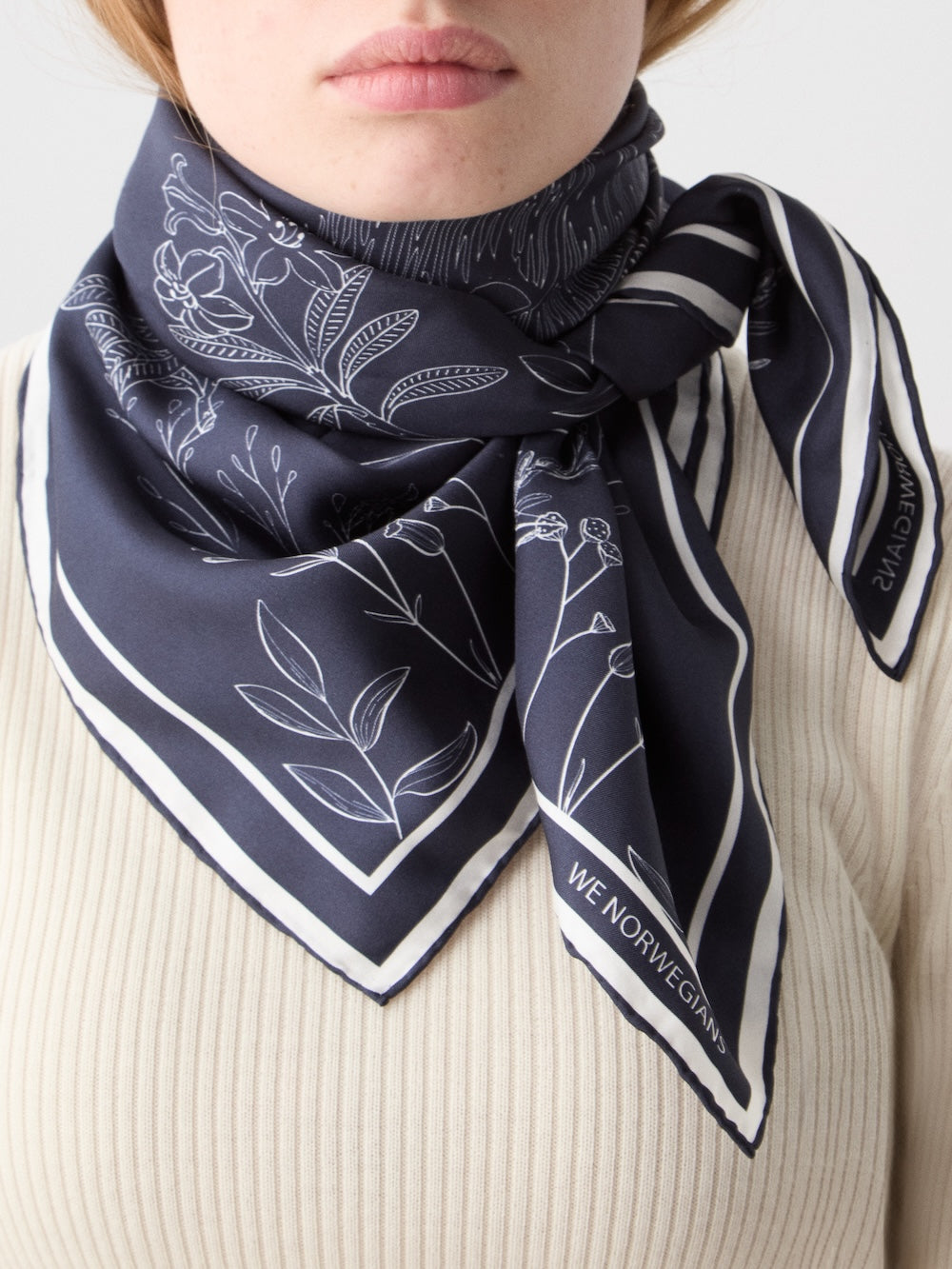 A person wears a cream ribbed top and the We Norwegians Alps Silk Scarf in dark navy with white floral line drawings, stripes, and "WE NORWEGIANS" text on one corner, neatly tied for a Scandinavian-inspired look.