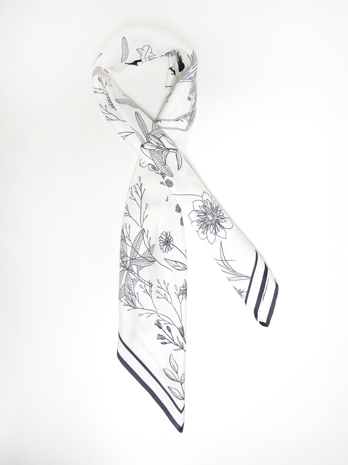 A pure silk white scarf with an intricate black floral pattern, neatly folded and hanging. The Alps Silk Scarf by We Norwegians features a variety of flowers and foliage, with a striped border along the edges. The background is plain white, highlighting the design of this exquisite silk scarf.