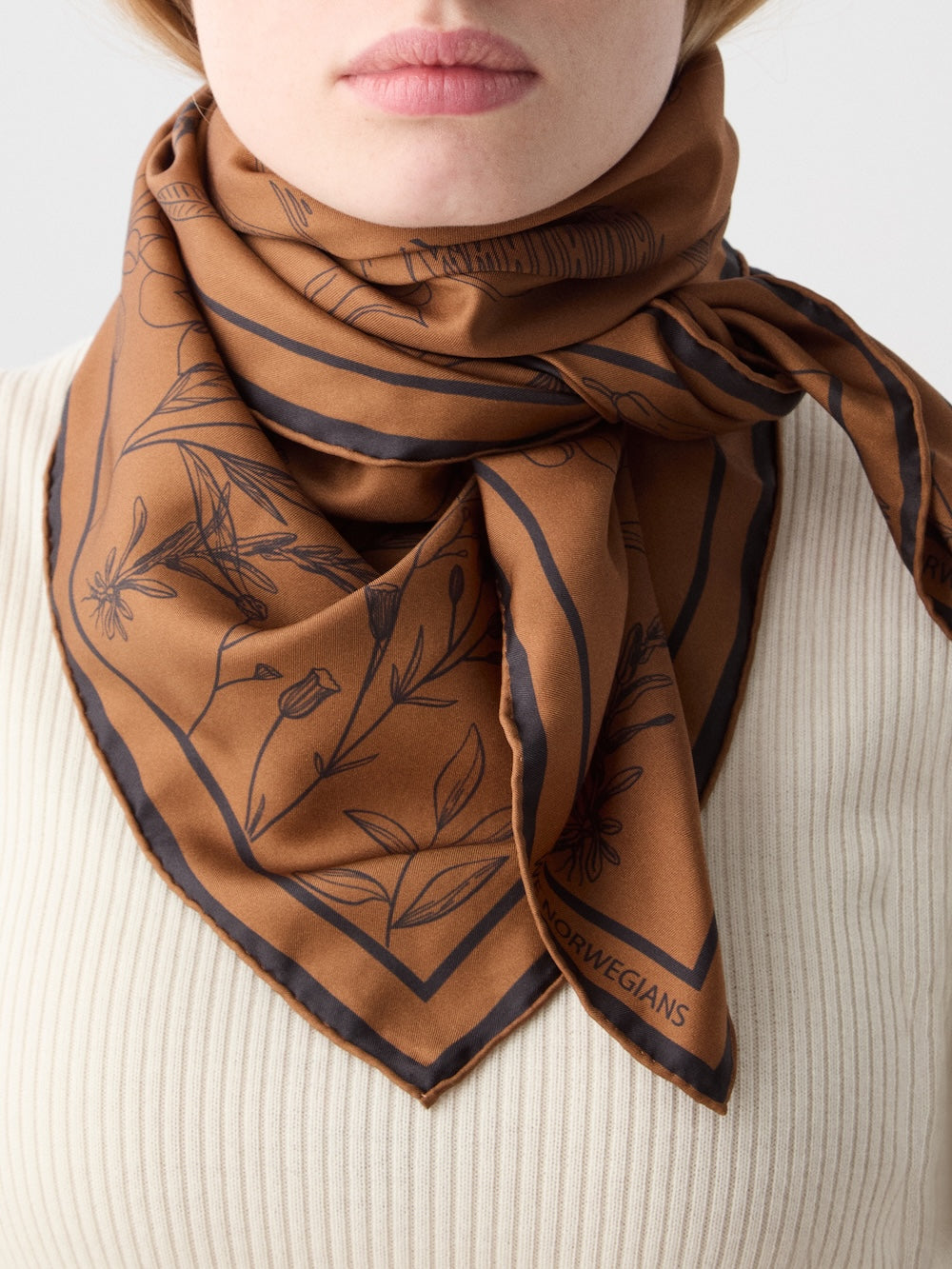 A person wears the Alps Silk Scarf by We Norwegians—brown silk with black floral outlines—tied around their neck over a cream ribbed top, showing only the lower face and neck for a chic après-ski style.