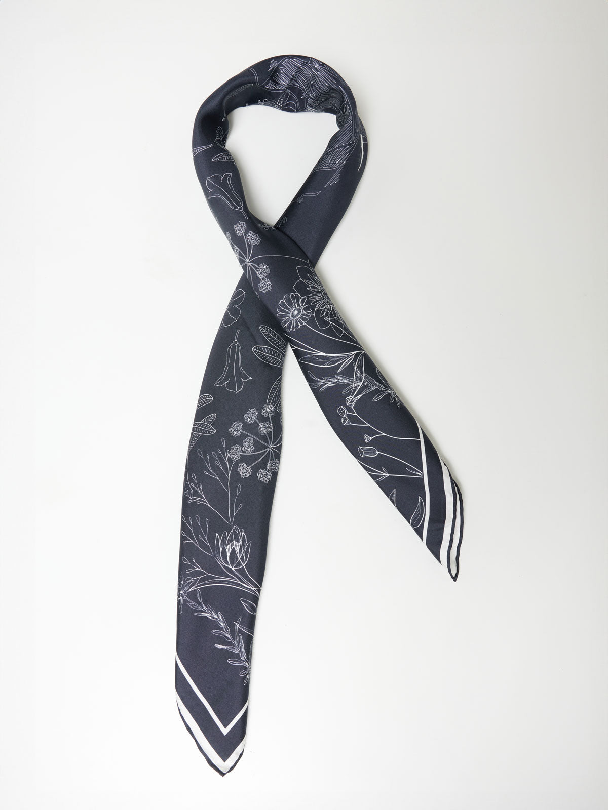 A sleek, dark grey Alps Silk Scarf by We Norwegians on a light background, featuring a subtle botanical pattern in white. Crafted from pure silk, the scarf has a thin white border near the edges, adding a touch of contrast to the elegant design.