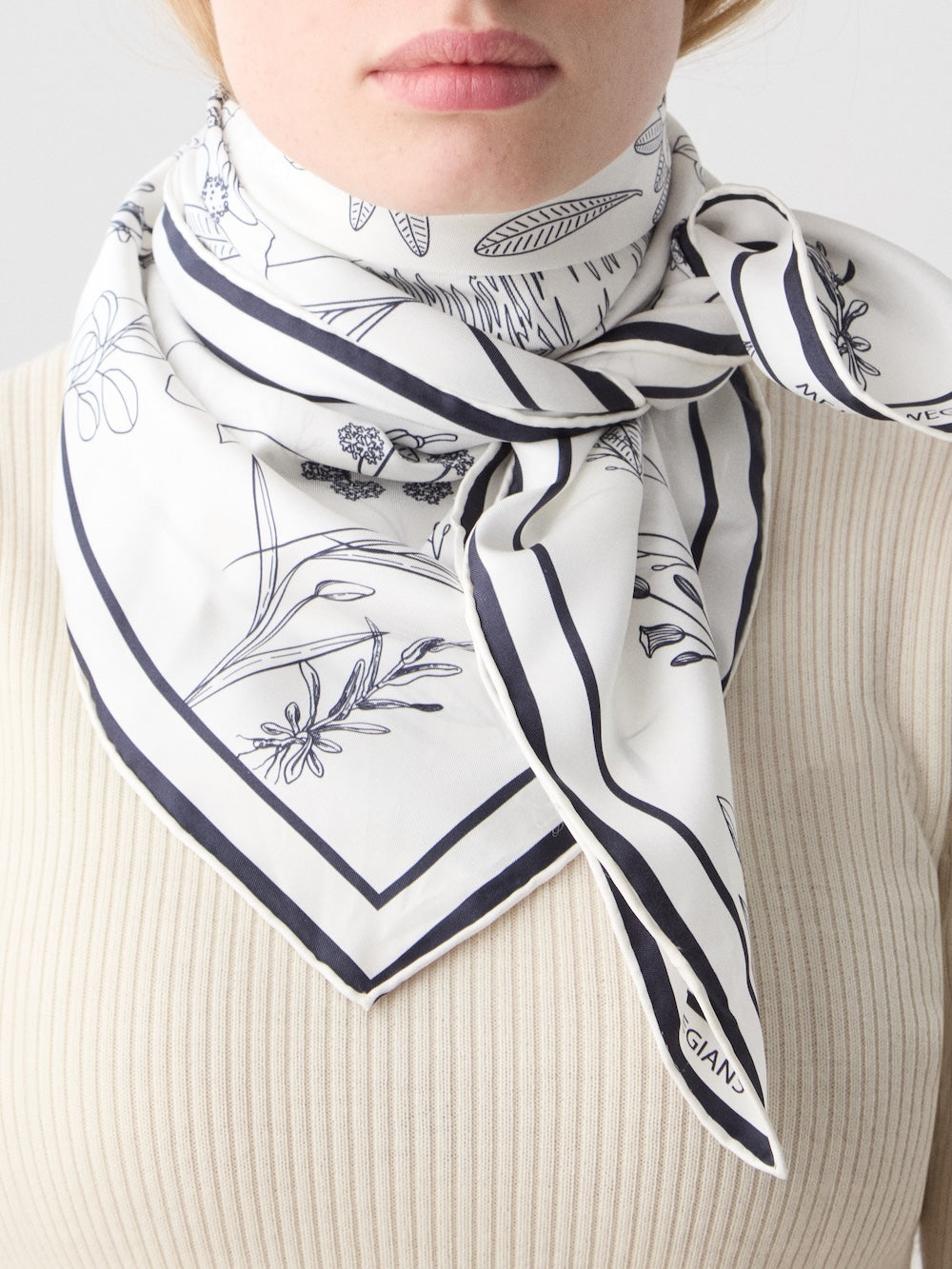 A woman wears a cream ribbed top and the Alps Silk Scarf by We Norwegians—a white silk scarf with black floral designs and borders, featuring intricate botanical illustrations for a Scandinavian-inspired elegance.