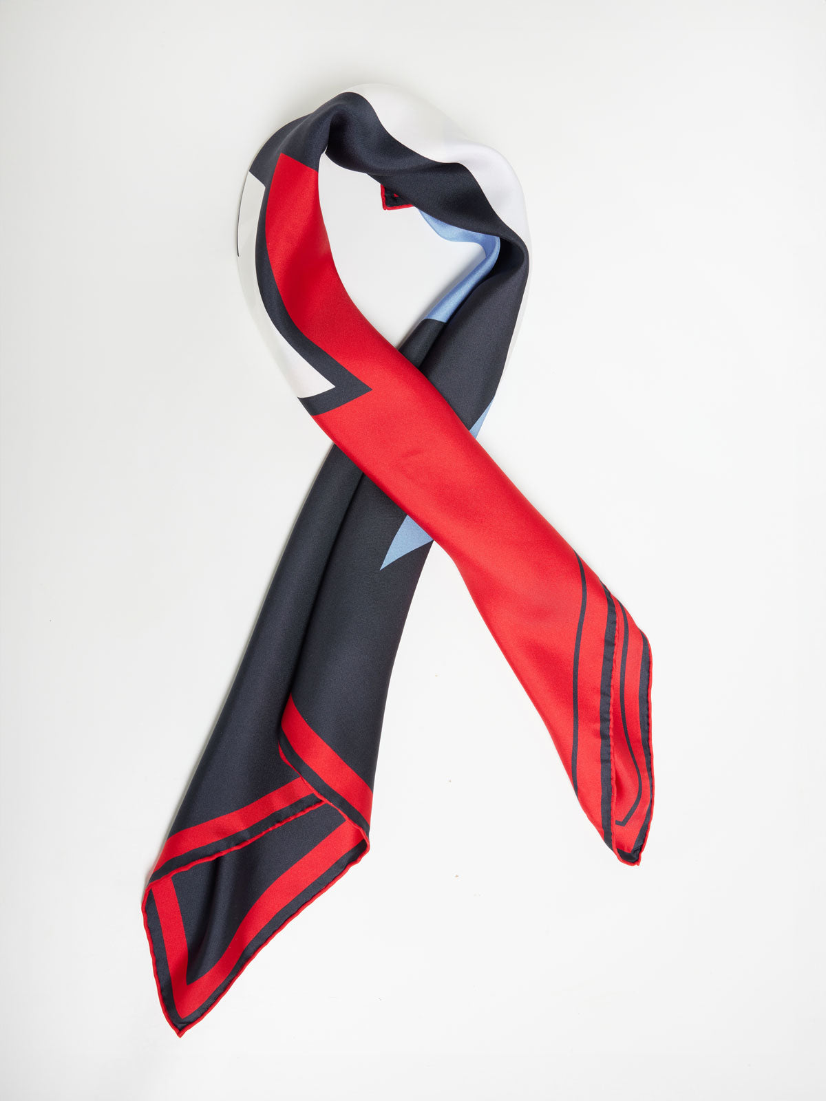 A We Norwegians Snowfun Silk Scarf, featuring a pure silk composition and a striking geometric design in black, red, and white. The scarf is loosely knotted into a loop against a snowy mountain backdrop, with bold red stripes and shapes that contrast beautifully with the surrounding black and white areas.