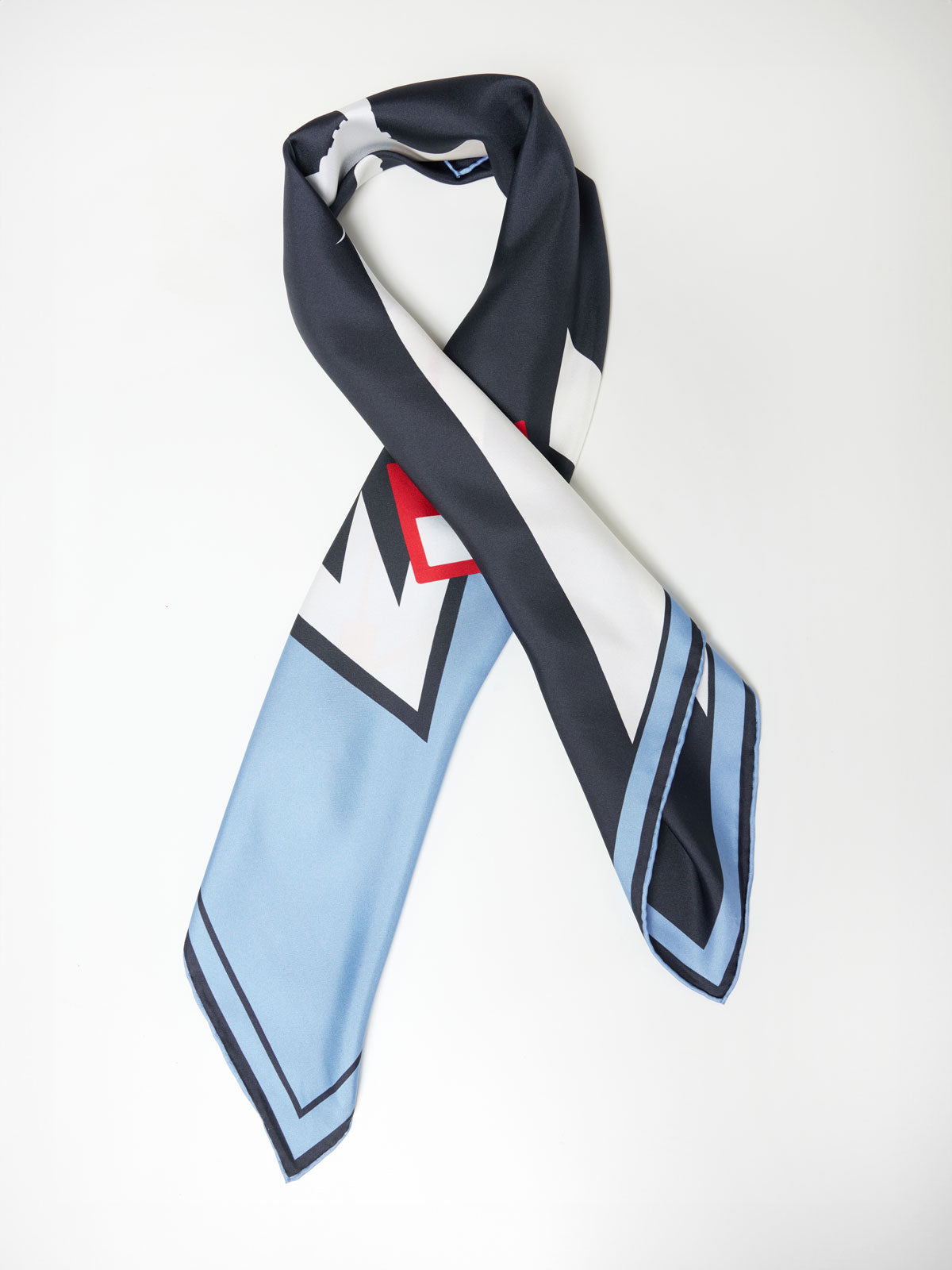 The Afterski Silk Scarf by We Norwegians is a stylish accessory with geometric patterns, showcasing a blend of black, white, blue, and red hues. The luxurious silk scarf is casually draped in a loop, highlighting its vibrant color blocks and crisp lines against a plain white background.