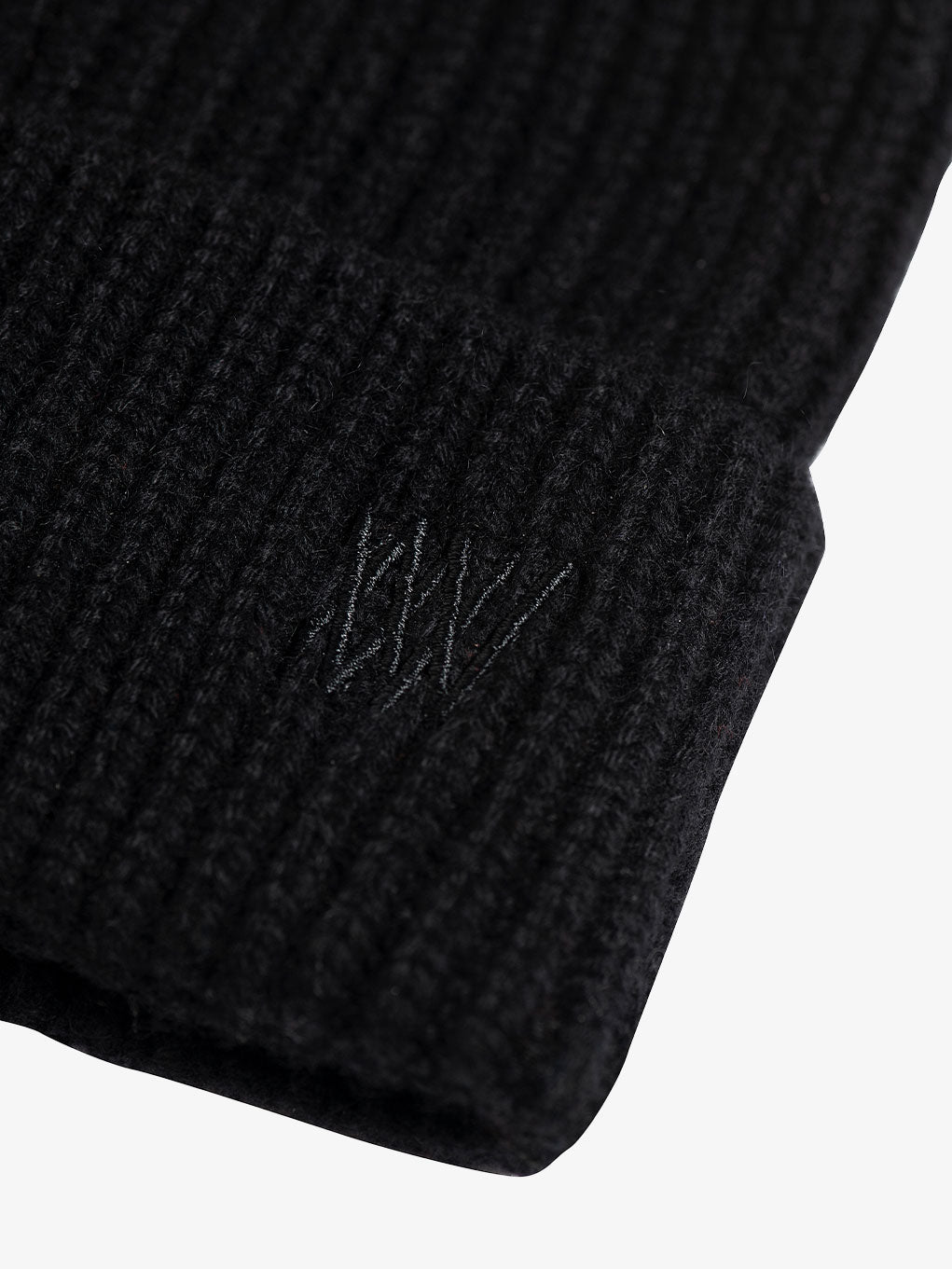 Close-up of the Blefjell Cashmere Hat by We Norwegians on a plain background. This black ribbed knit beanie features subtle tonal embroidery and an adjustable cuff on the edge.