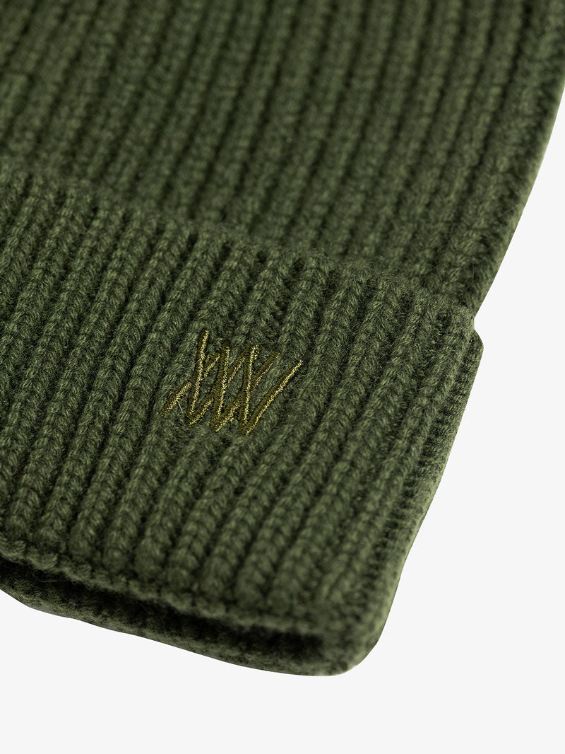 A close-up of the Blefjell Hat by We Norwegians showcases its green ribbed knit design, complete with tonal embroidery and an adjustable cuff. The logo is intricately embroidered in a slightly darker green thread, emphasizing the texture of the fabric and the subtle stitched design.