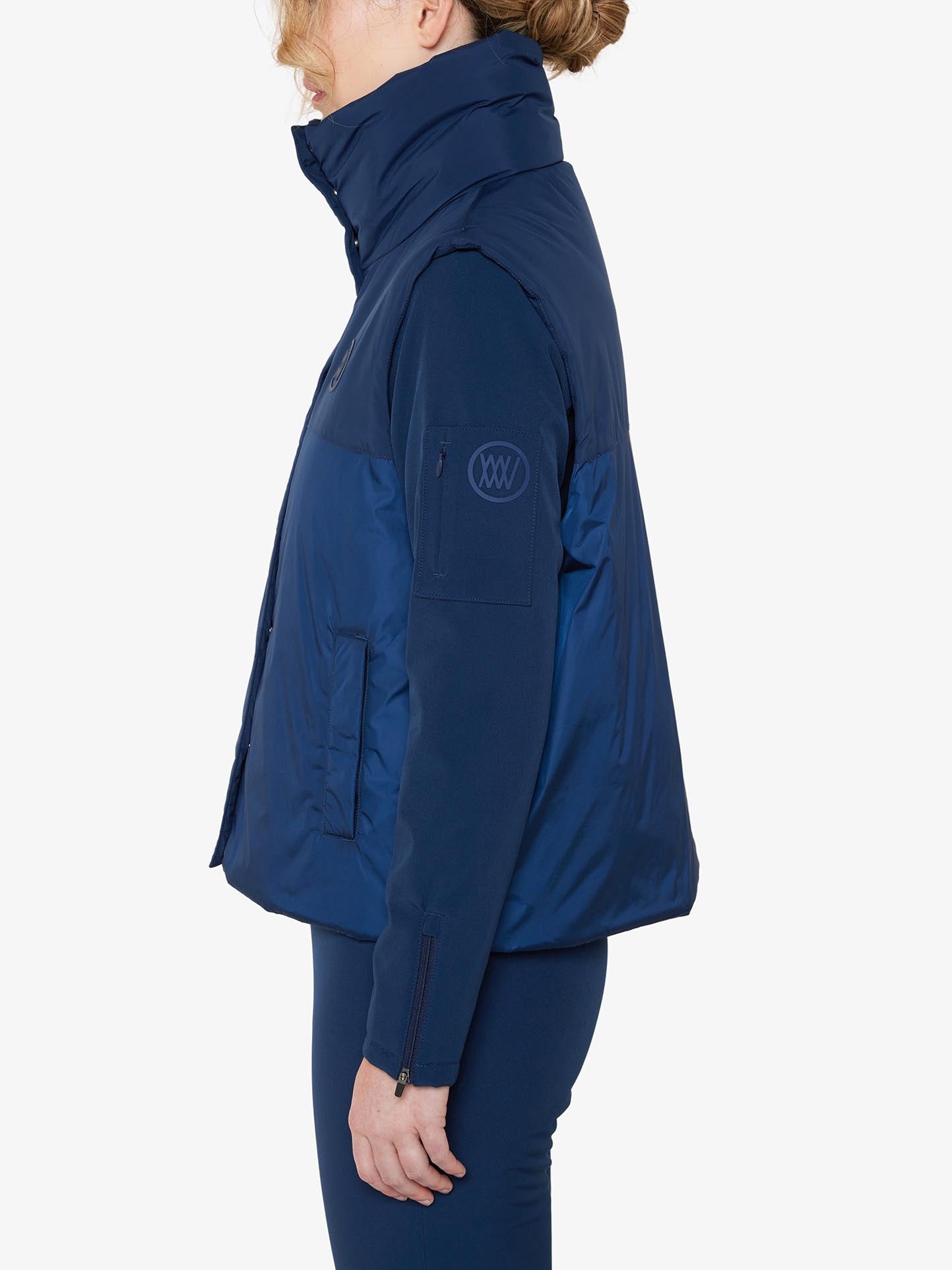 A functional fashionista is shown from the side, wearing the We Norwegians Filefjell Vest Women in navy blue. The high-collared, long-sleeve puffer jacket is made of water-repellent fabric and features front pockets along with a small circular logo on the upper arm. She pairs it with dark blue pants against a plain white background.