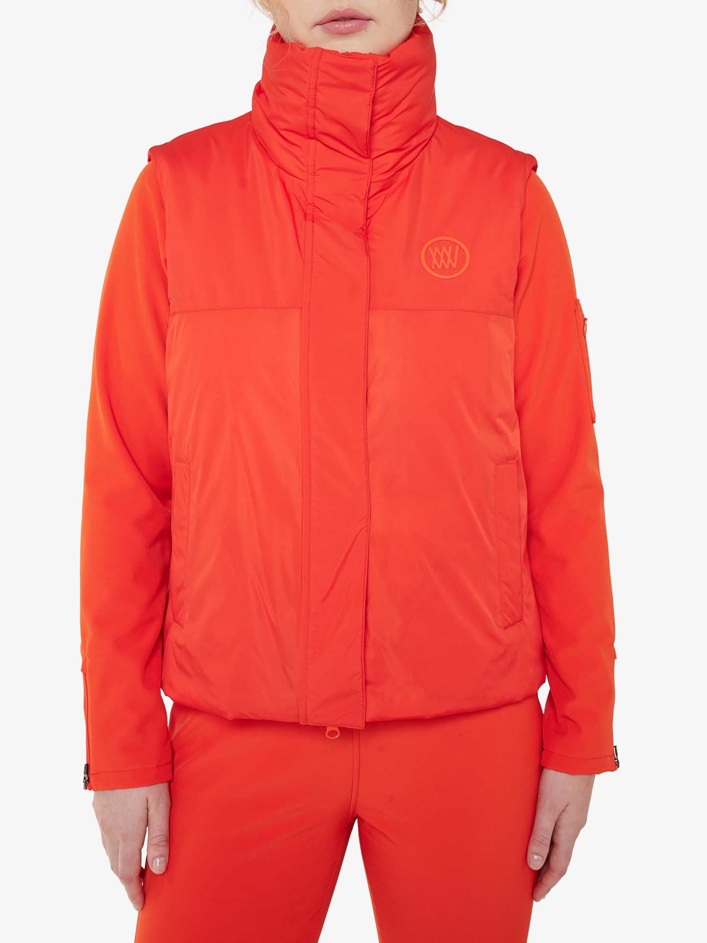 A functional fashionista is wearing the bright orange Filefjell Vest for Women by We Norwegians. With a high neck and logo on the left chest, this padded jacket is made from water-repellent fabric and features side pockets. The outfit includes matching orange pants, creating a clean aesthetic that stands out against the plain white background.