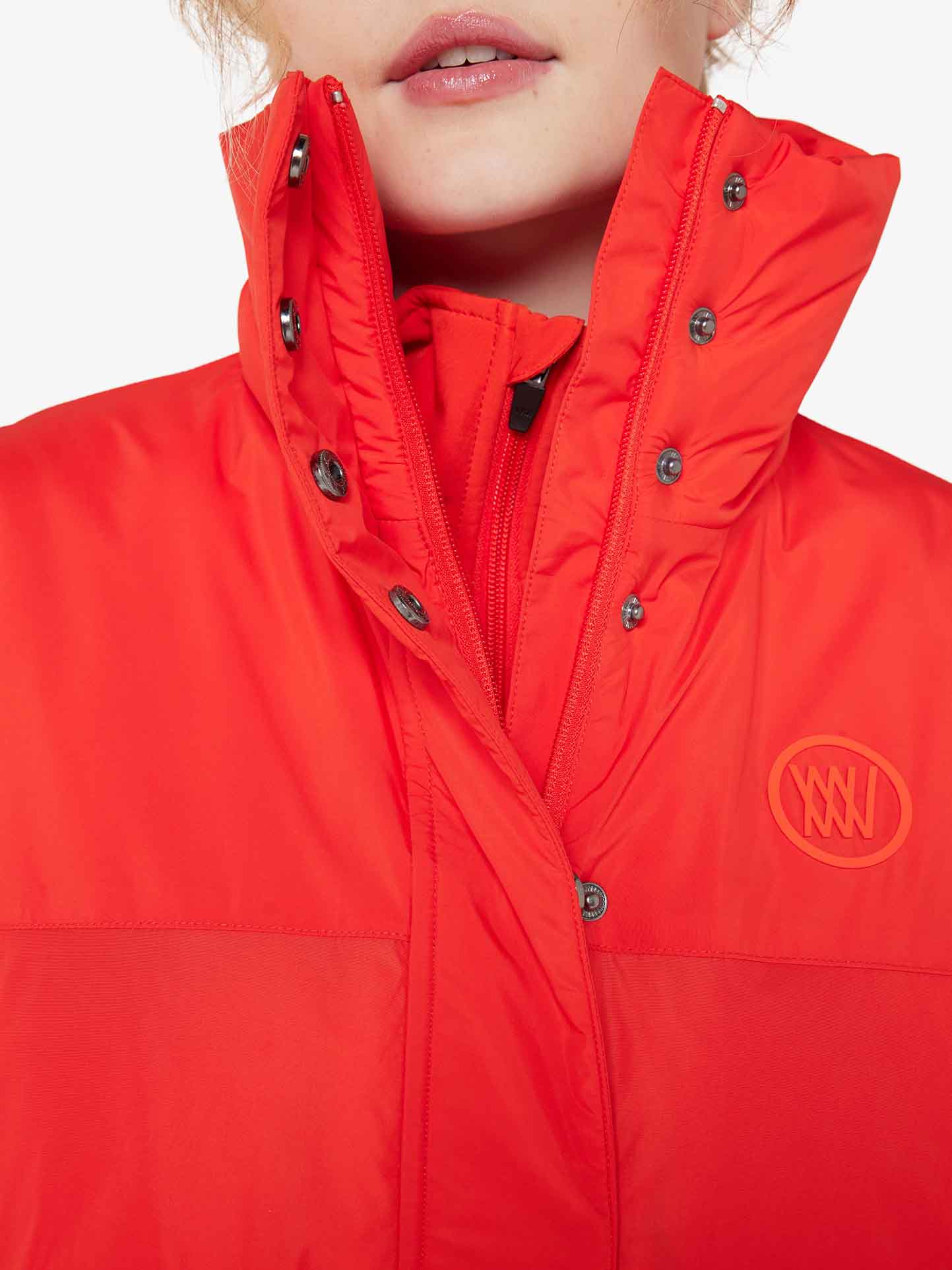 Close-up of a person wearing the We Norwegians Filefjell Vest Women, a bright red winter jacket with wool padding and a high collar. This functional fashionista choice features a zipper and black snaps on the collar. A circular logo with wavy lines is visible on the chest, with the person's lips slightly visible at the top of the image.