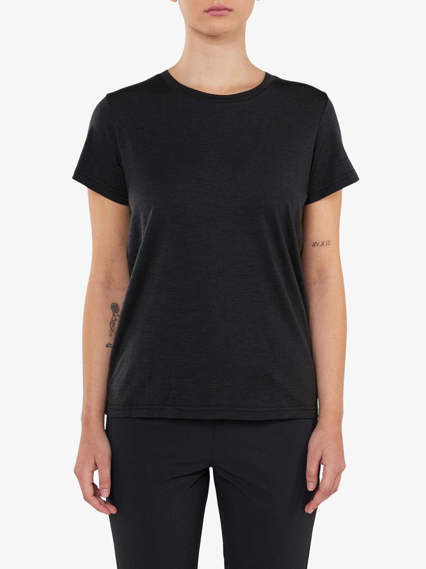 A person stands against a plain background, wearing a fitted black Berg Favorite Tee Women by We Norwegians and black pants. They have tattoos on both forearms: a simple design on the left and Roman numerals on the right. Despite their relaxed arms by their sides, the no-itch Merino wool fabric ensures a comfortable fit.