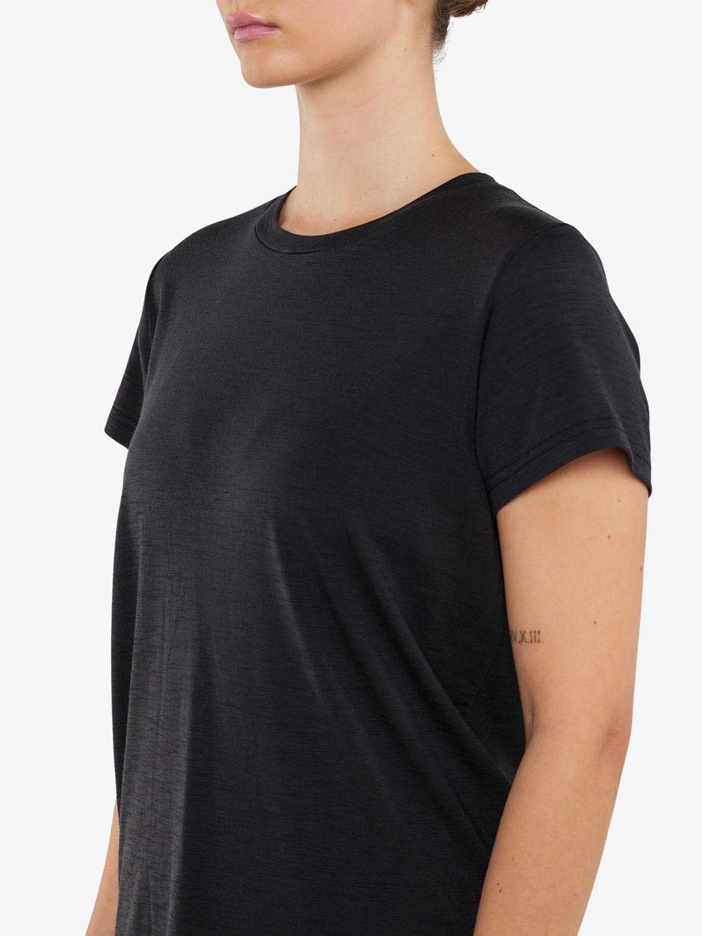 A person wearing a plain black Berg Favorite Tee Women from We Norwegians is shown from the shoulders to mid-torso, facing slightly to the right. Only part of their face and arms are visible. A small tattoo with Roman numerals graces the inner side of their right upper arm, highlighting the tee's comfortable fit and no-itch merino wool fabric.