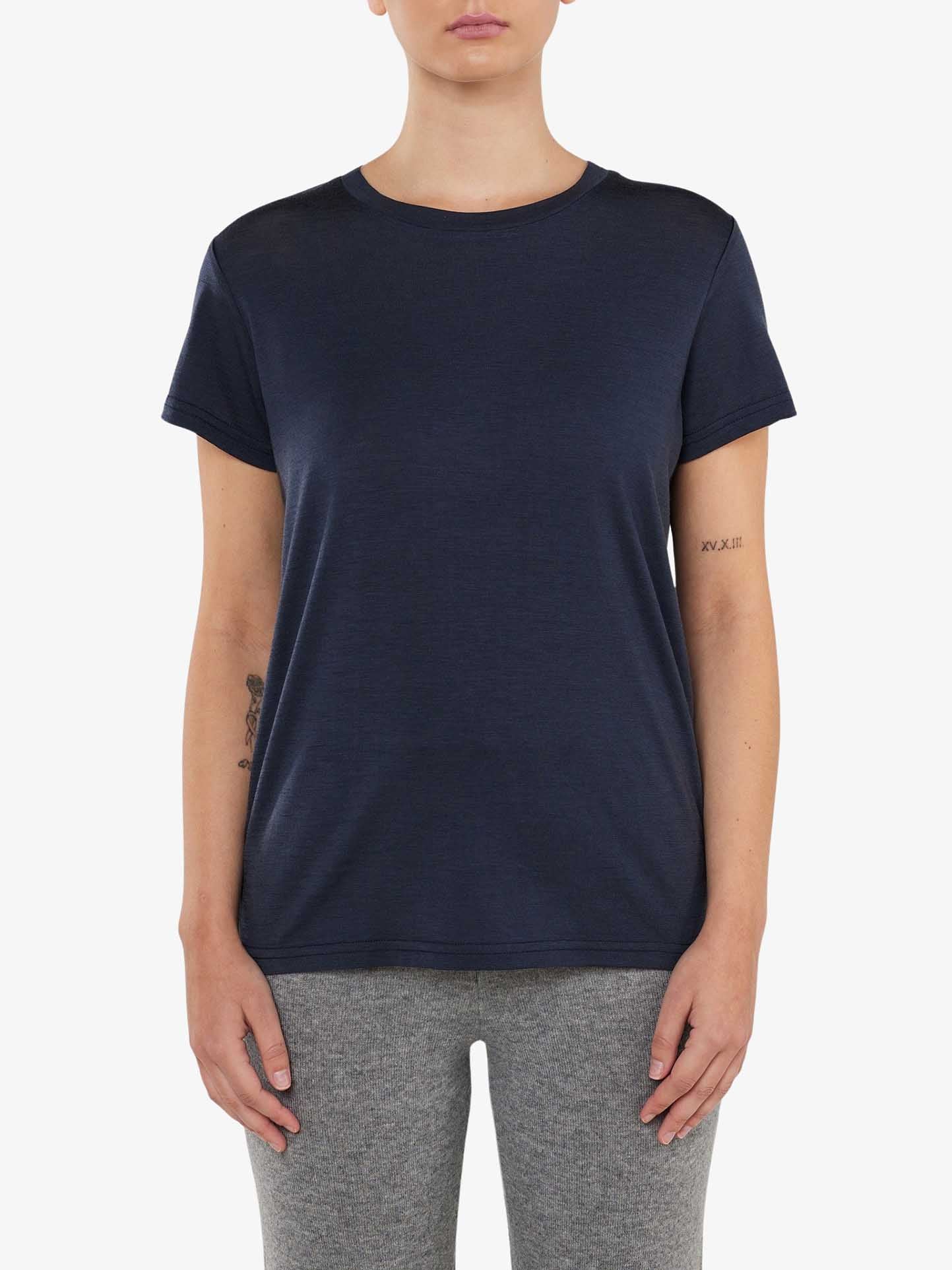 A person standing and facing forward, wearing a comfortable fit dark blue "Berg Favorite Tee Women" by We Norwegians and light grey pants. They have a small tattoo on their left forearm and one on their right inner bicep. The background is plain white.