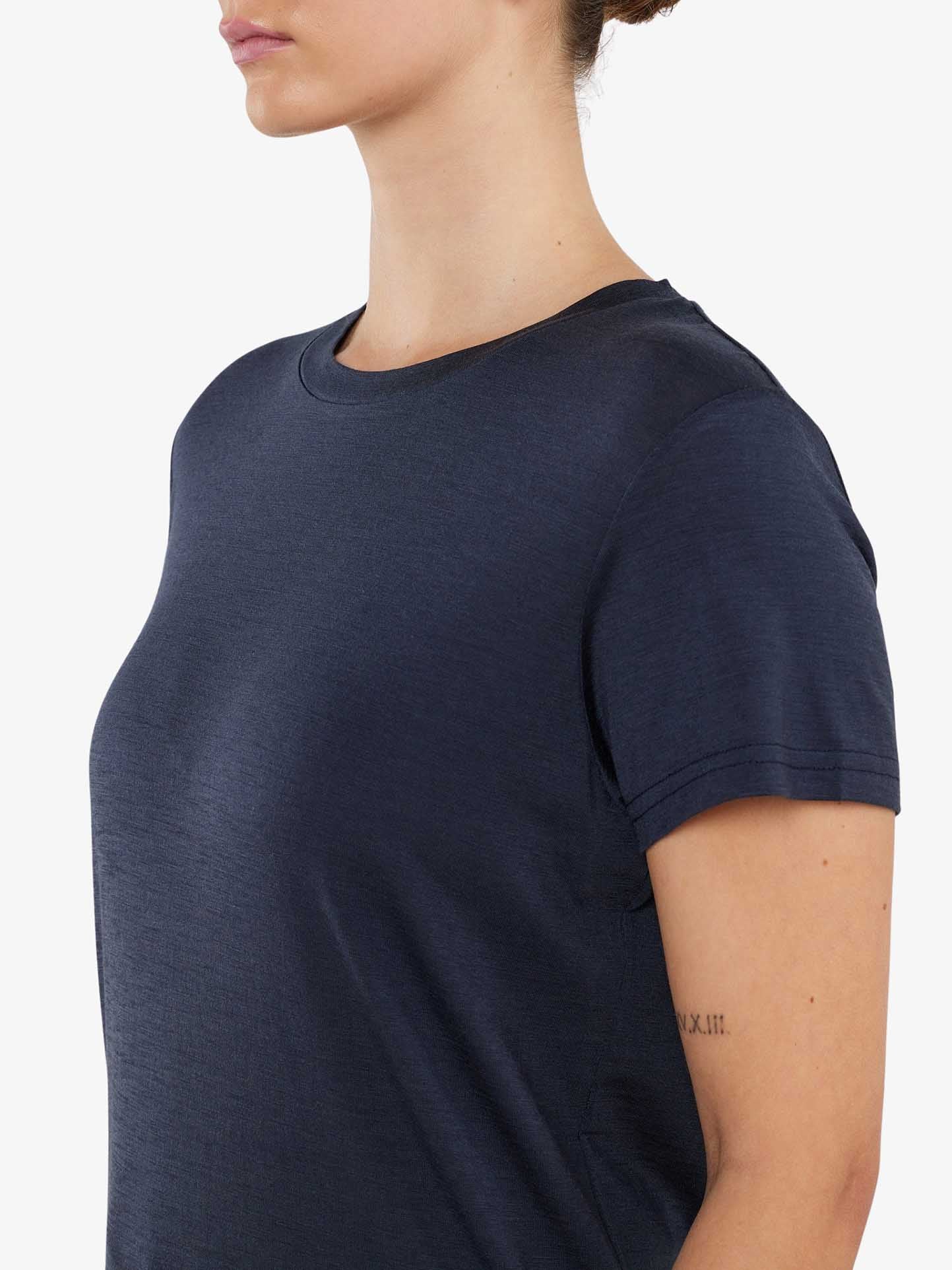 A person wearing the Berg Favorite Tee Women by We Norwegians is shown from the side, facing left. They have a subtle tattoo of Roman numerals on their upper arm. Their hair is tied back in a neat bun, and the background is a solid, light color. The navy blue short-sleeve merino wool tee provides a comfortable fit with no-itch fabric.