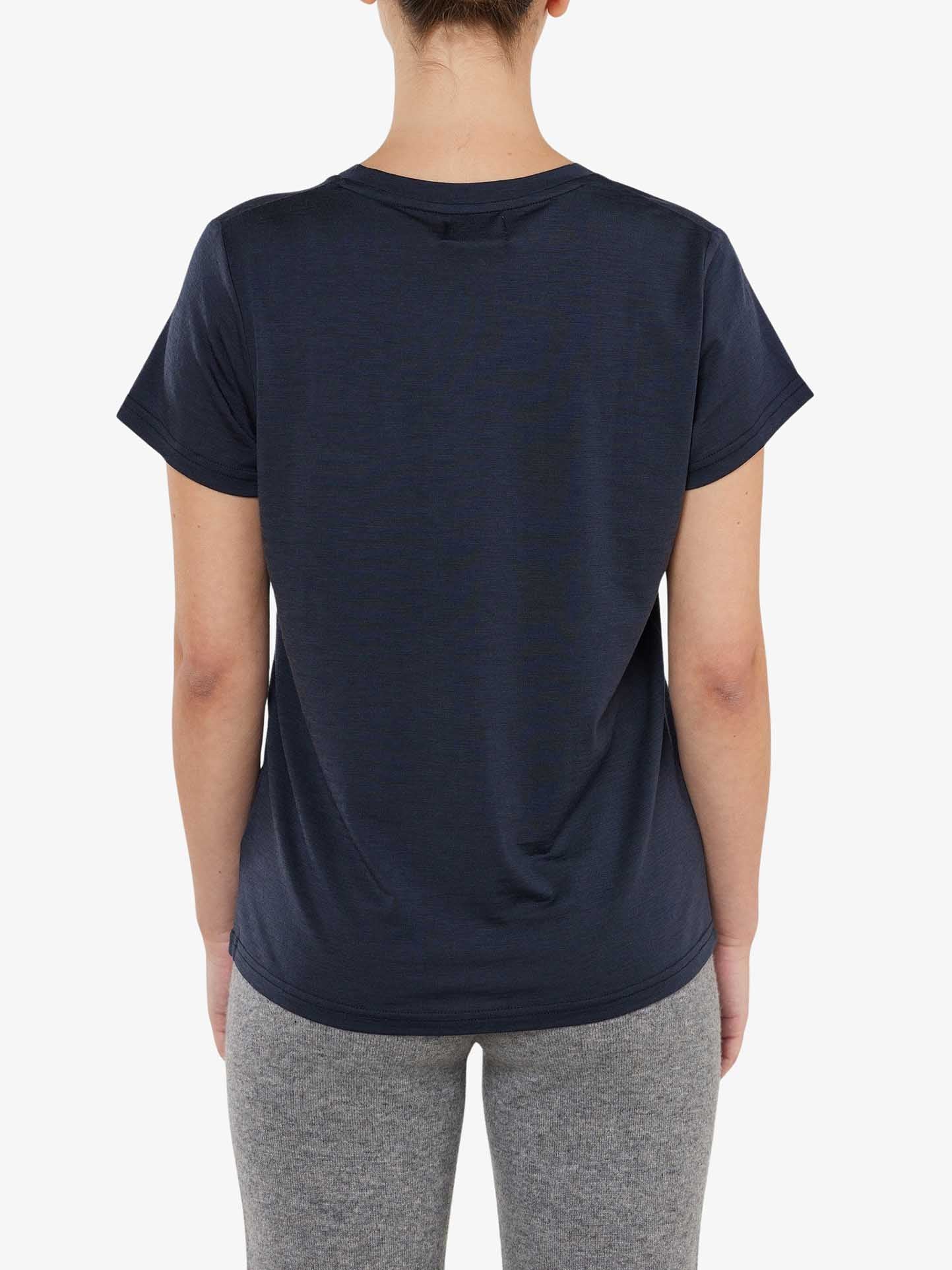 Rear view of a person dressed in a Berg Favorite Tee Women from We Norwegians, a dark navy short-sleeve merino wool t-shirt paired with light grey pants. The person stands against a plain white background, showcasing the comfortable fit and no-itch fabric of the tee.