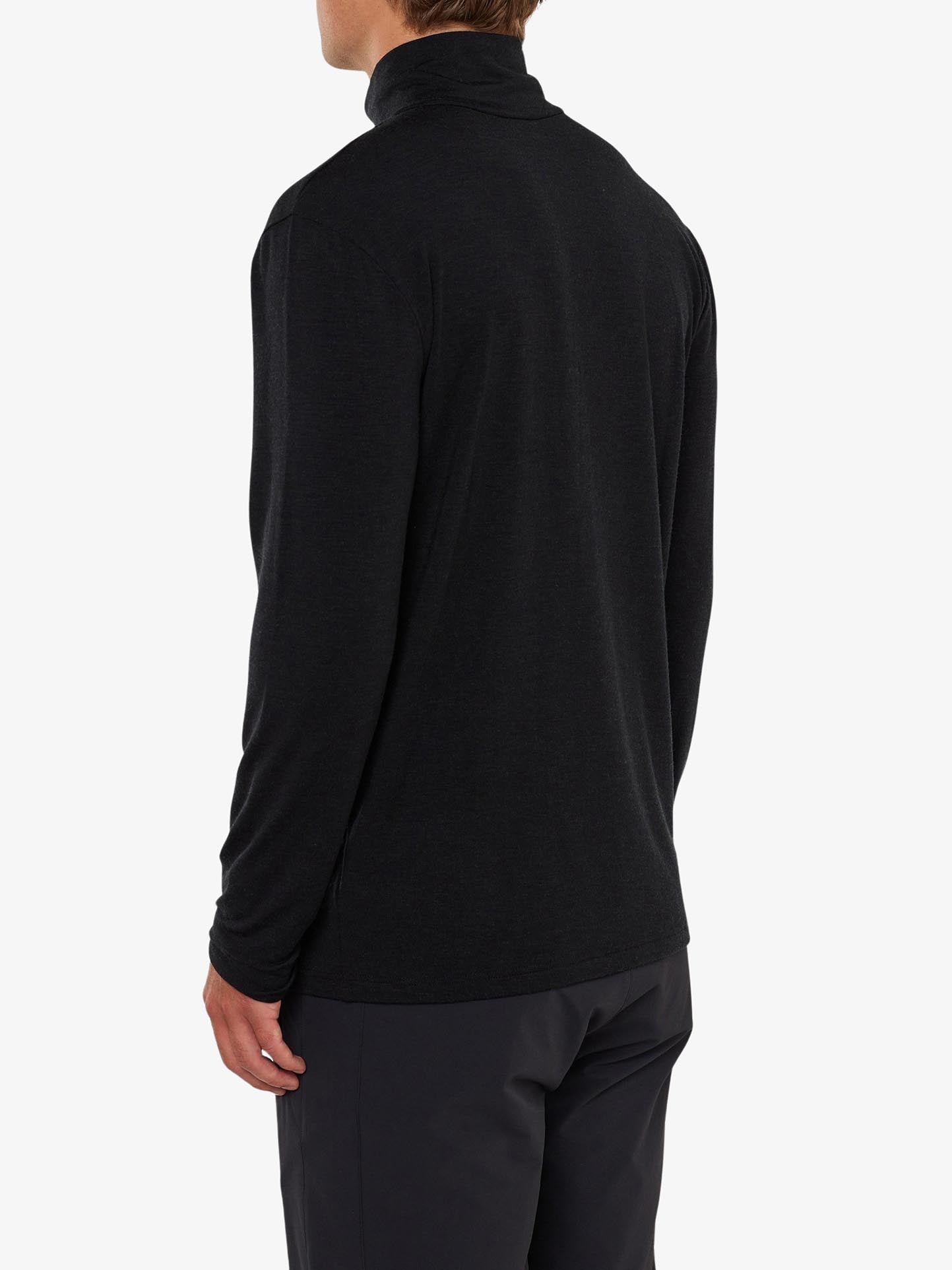 A person is shown from the back, wearing the We Norwegians Hygge ZipUp Men in black paired with dark pants. The background is plain white, emphasizing the luxurious base layer made of fine merino wool.