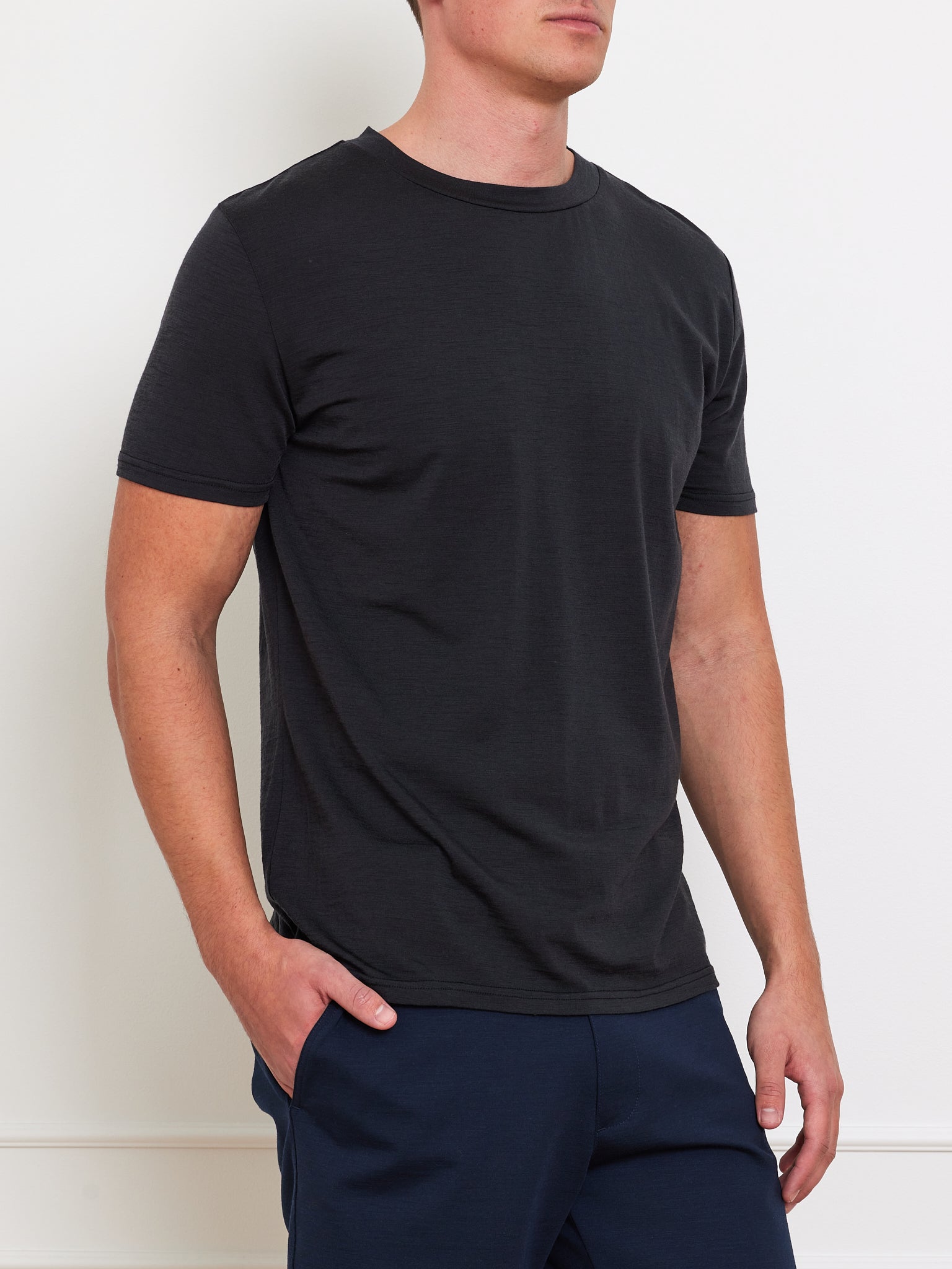 A man embodies classic style in a Berg Tee Men from We Norwegians, crafted from cooling fabric. He pairs it with dark blue pants, set against a white backdrop. With effortless flair, he casually places one hand in his pocket and faces right.