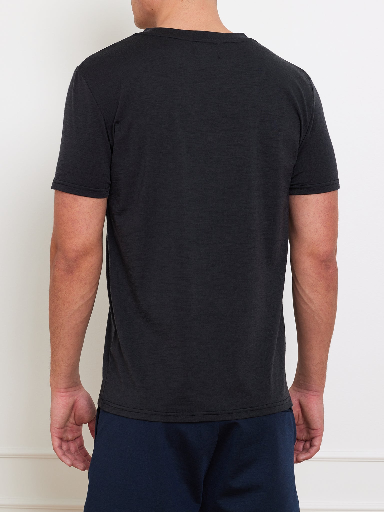 A person wearing a We Norwegians Berg Tee Men in black, made of cooling fabric, and dark pants stands with their back to the camera. The plain white wall complements their versatile wardrobe choice, perfect for any season.