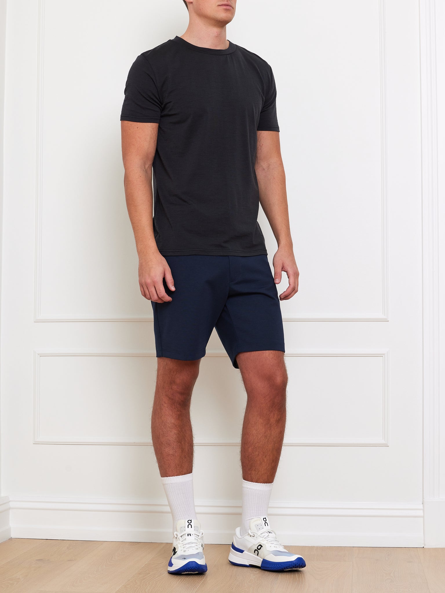 A person in a Berg Tee Men by We Norwegians and navy blue cooling fabric shorts, complemented by white socks and white-blue sneakers, stands against a white wall. Their face is hidden, while their outfit pairs perfectly with the room's light wooden floor.