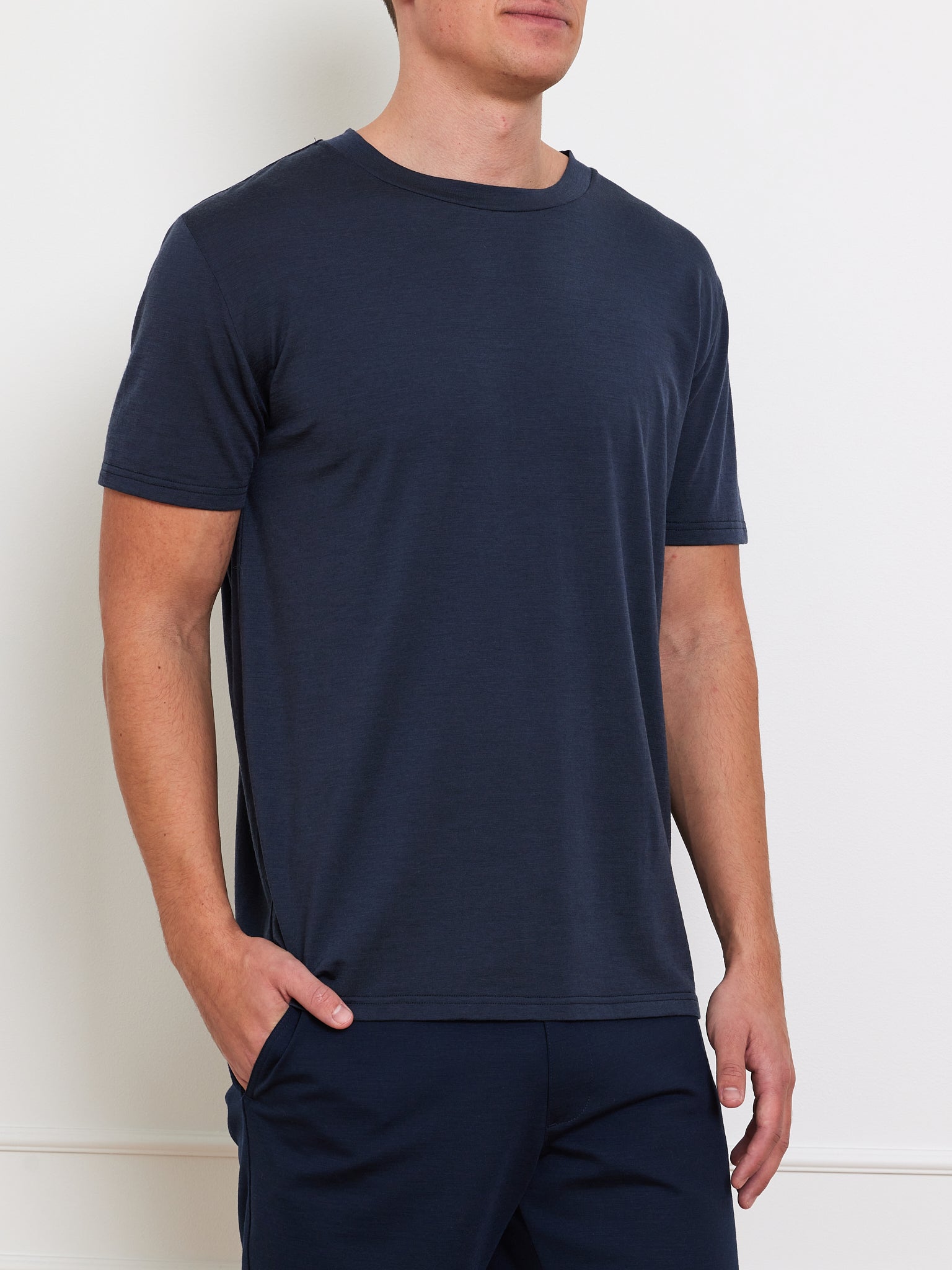 A man stands against a white wall, stylishly clad in the Berg Tee Men by We Norwegians. The cooling Merino blend fabric of his plain dark blue T-shirt and pants makes it an appealing wardrobe staple, as he casually tucks one hand in his pocket.