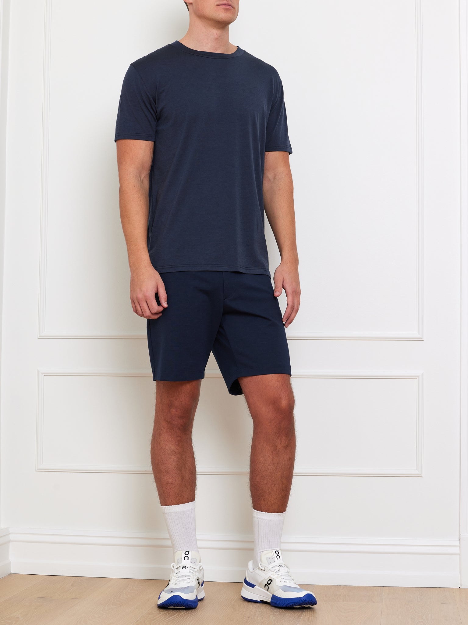 A person in a Berg Tee Men by We Norwegians and shorts stands against a white wall with wooden flooring, clad in white socks and blue-white cooling fabric sneakers. The head is not visible.