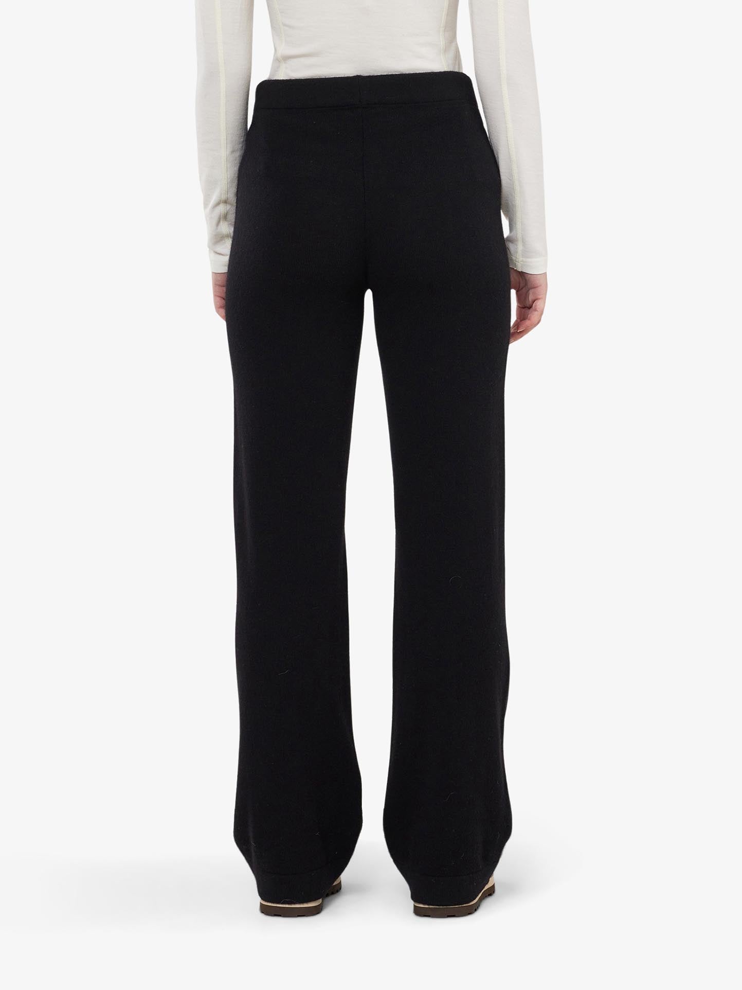 The image highlights a back view of We Norwegians' Blefjell Cashmere Pant Women in black, paired with a long-sleeved white top. The outfit stands elegantly against a plain white background.