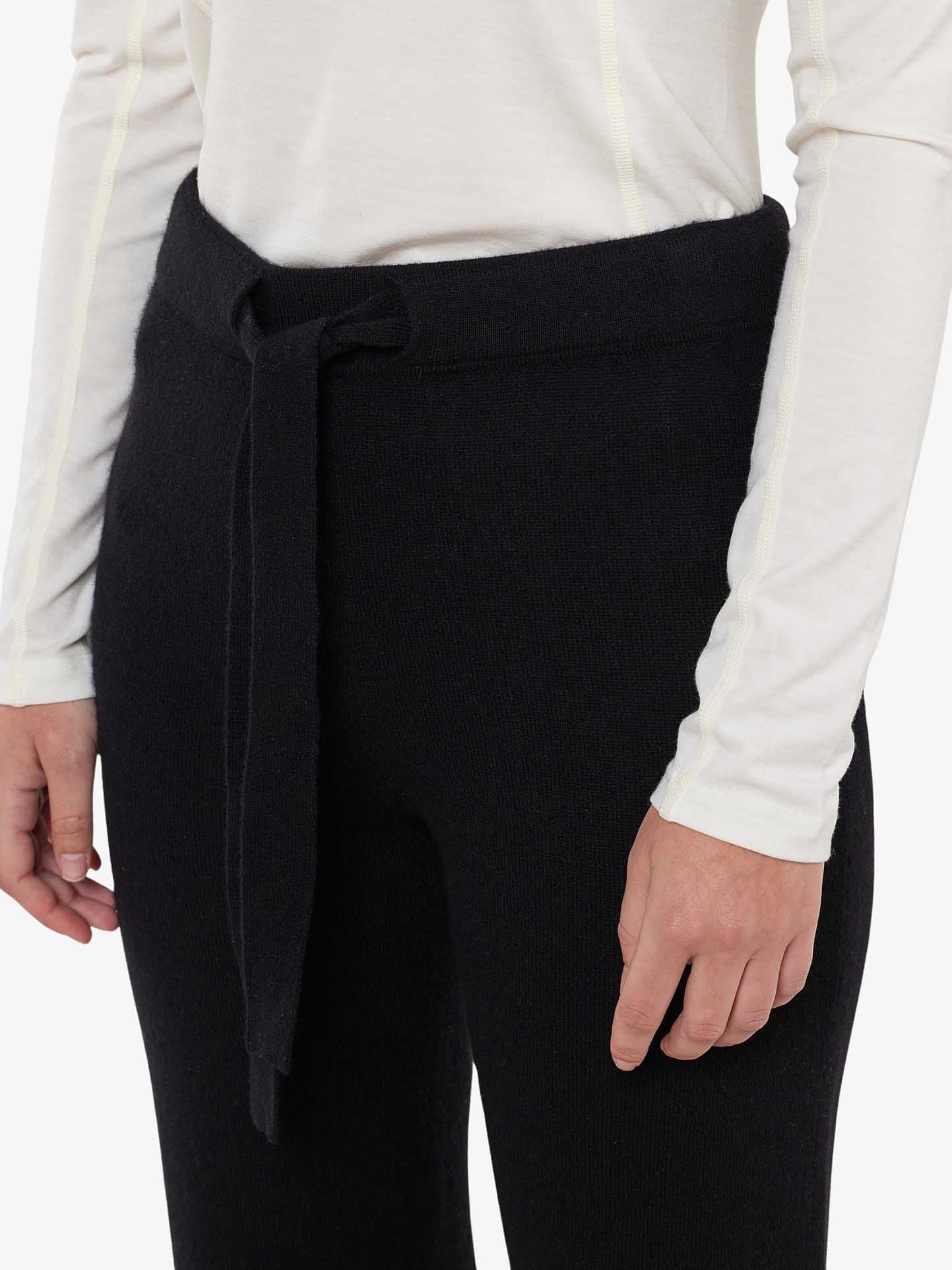 A person wears a long-sleeved white shirt paired with the Blefjell Cashmere Pant for Women by We Norwegians, featuring high-waisted, wide-leg black pants with a fabric belt tie. The cropped shot focuses on the torso and hands, all set against a plain backdrop from the elegant Blefjell Collection.