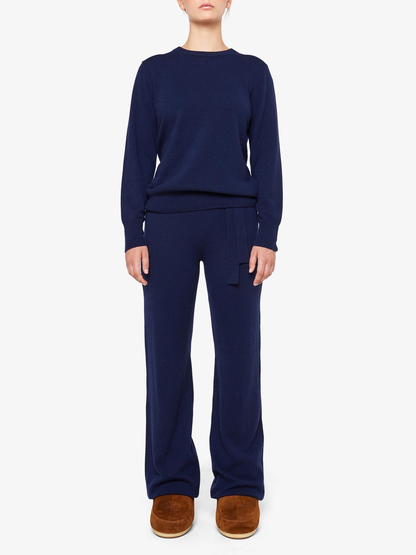 A person stands facing forward, dressed in a navy blue 100% Cashmere long-sleeve top and matching Blefjell Cashmere Pant from We Norwegians. Brown slippers complete the look against a plain white background.