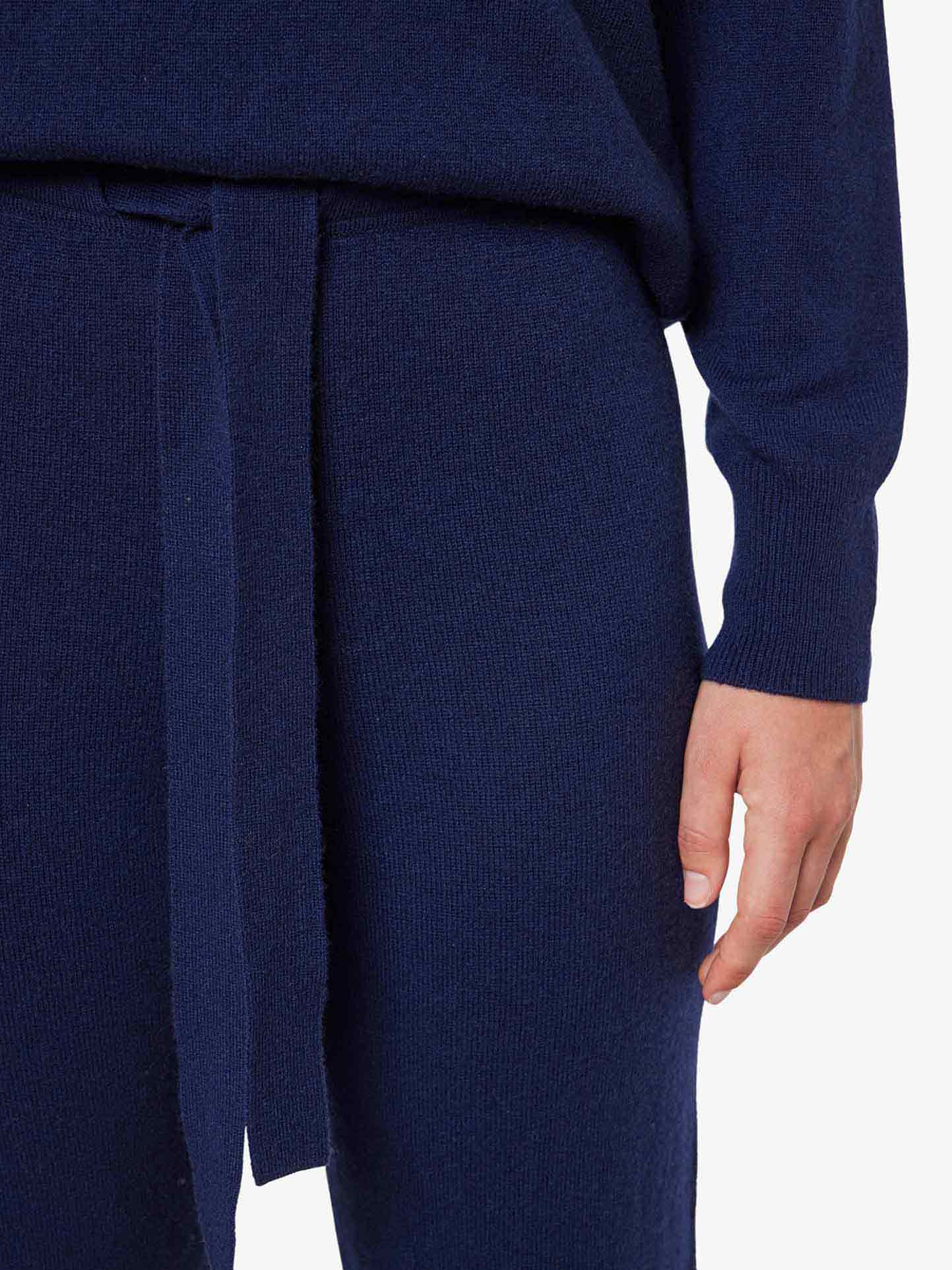 A person is dressed in a dark blue knit outfit from the Blefjell Collection by We Norwegians, showcasing the Blefjell Cashmere Pant Women with wide legs and a tie belt, along with a long sleeve top. Only the lower torso, right arm, and hand are visible against a plain white background.