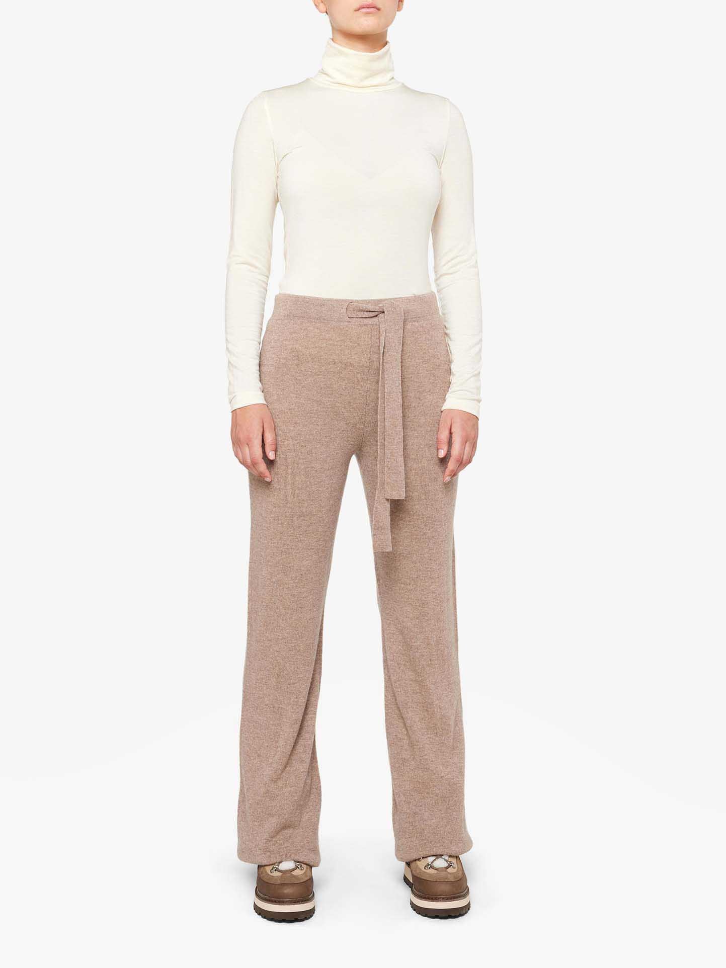 A person showcases the Blefjell Collection by We Norwegians, wearing a cream 100% cashmere turtleneck sweater paired with the Blefjell Cashmere Pant Women in beige, featuring a textured wide-leg design and tied waistband, complemented by brown shoes against a plain white background.