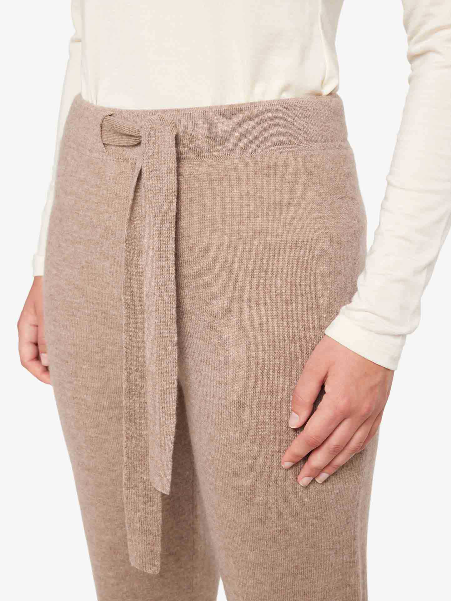 A person is seen wearing the We Norwegians Blefjell Cashmere Pant Women in beige, crafted from 100% cashmere and featuring a matching fabric belt, paired with a white long-sleeve top. The image highlights the lower torso, emphasizing the luxurious texture and fit of these pants from the Blefjell Collection.