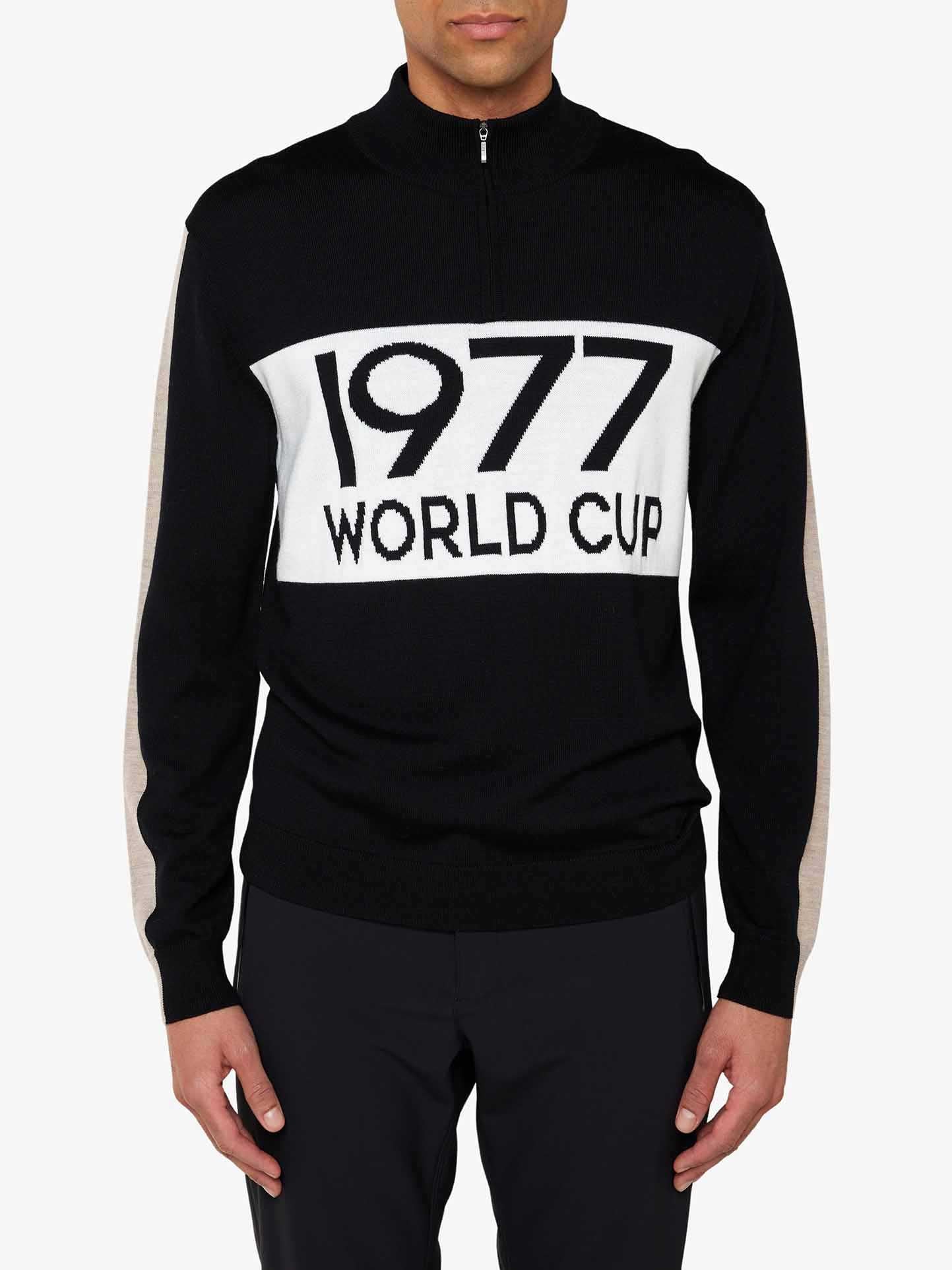 A person is wearing the We Norwegians World Cup Zipup Men, a black long-sleeve sweater featuring a white panel across the chest with "1977 WORLD CUP" in large, bold letters. Embracing 70's ski fashion, this sweater includes a half-zip collar and contrasting beige stripes down the sleeves. The person is also sporting black pants.