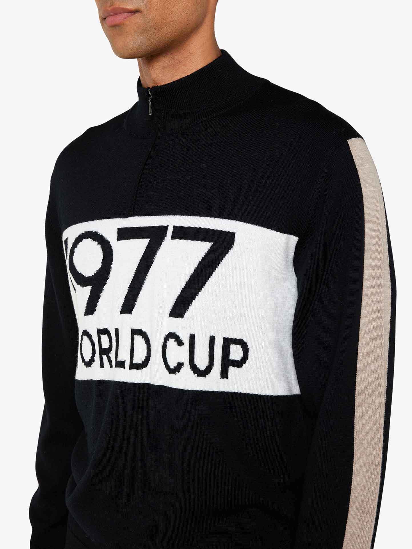 Someone is highlighting a We Norwegians sweater, the 1977 World Cup Zipup Men. This black high-collared zip-up features a bold white "1977 WORLD CUP" stripe on the chest and a beige sleeve stripe, reflecting the vibrant 70's ski fashion akin to Ingemar Stenmark's iconic style.