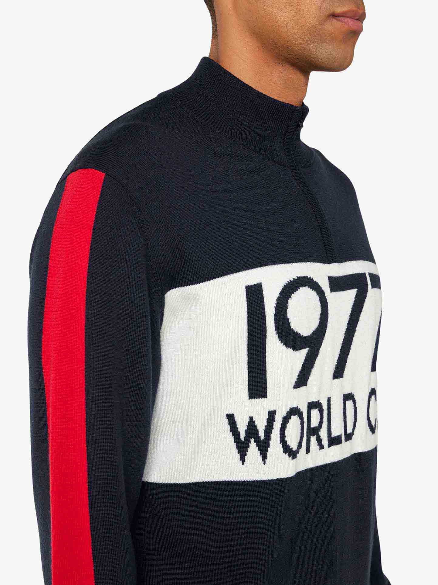 Wearing a We Norwegians 1977 World Cup Zipup Men sweater, featuring a zip-up collar with white and red sleeve stripes, and "1977 WORLD CUP" on the front, reminiscent of Ingemar Stenmark's era. The background is light gray.