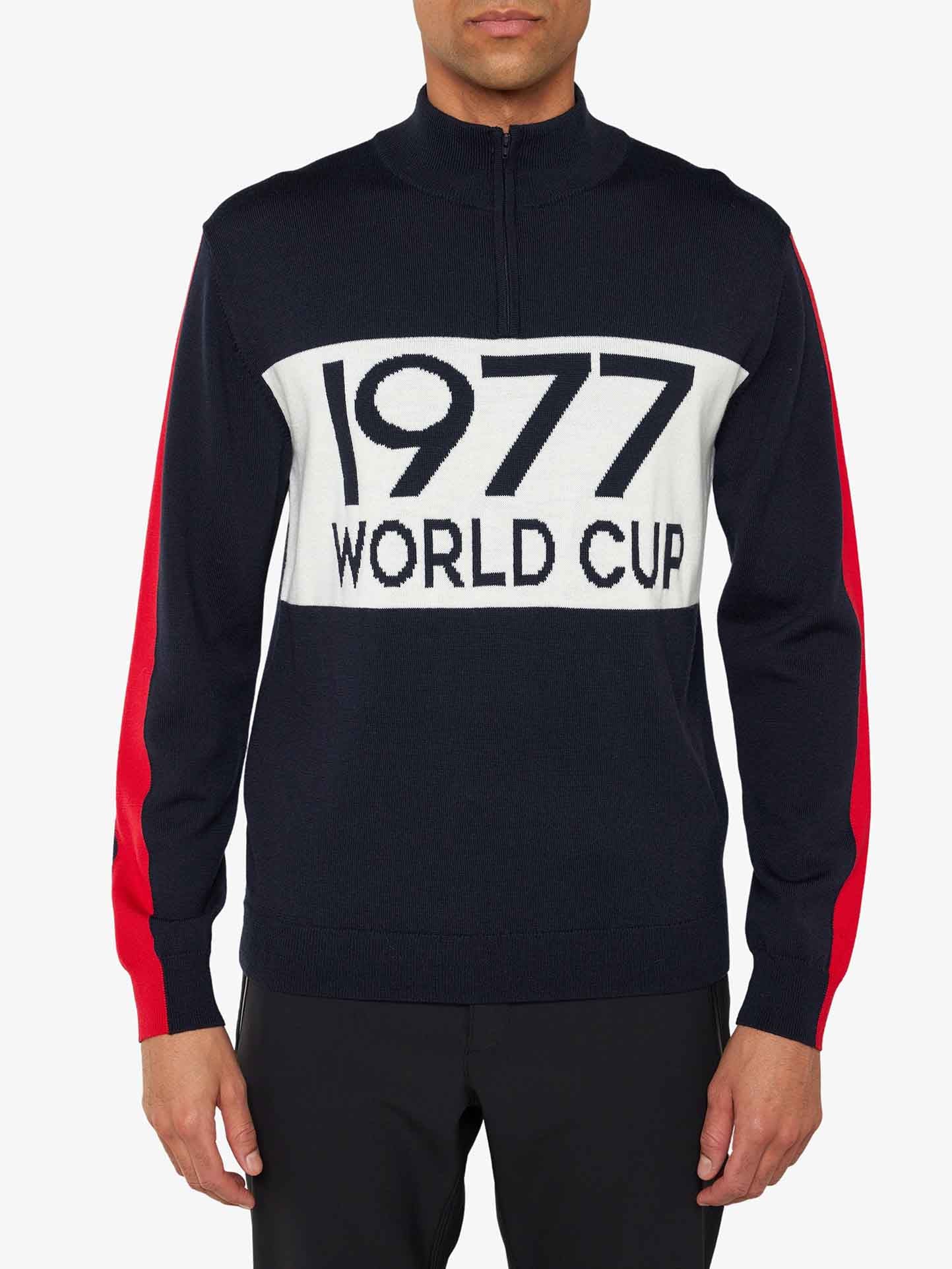 A man channels 70's ski fashion in the "1977 World Cup Zipup Men" by We Norwegians, a black sweater with a white "1977 WORLD CUP" chest panel. It features red sleeve stripes and a half-zip collar, reminiscent of Ingemar Stenmark's era, paired effortlessly with black pants.