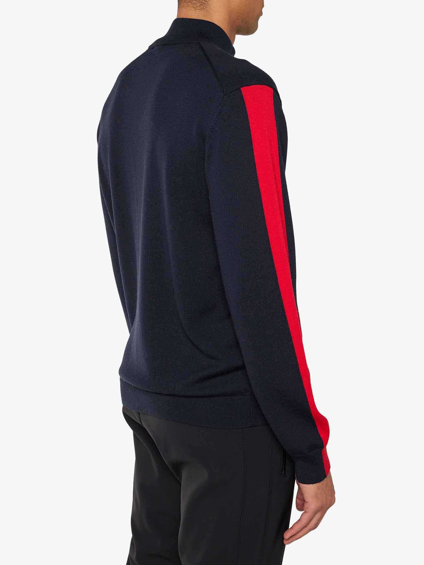 A person is wearing a We Norwegians World Cup Zipup Men in dark navy, featuring bold red stripes running down the length of each sleeve. They are facing away from the camera and paired it with black pants, evoking classic '70s ski fashion against a plain white background.