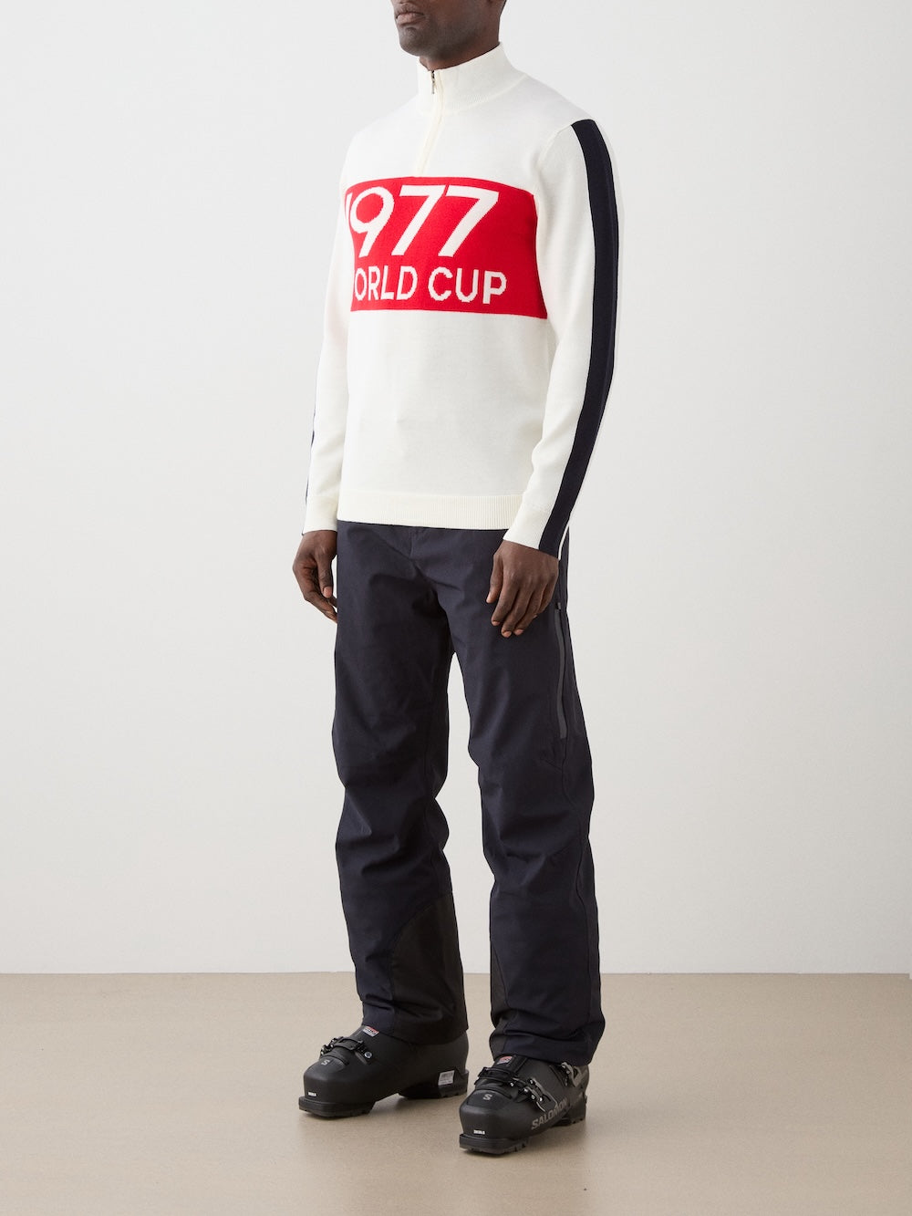 A person stands indoors against a plain light background, wearing the We Norwegians 1977 World Cup Zipup Men sweater, black pants, and black ski boots—showcasing classic alpine style.