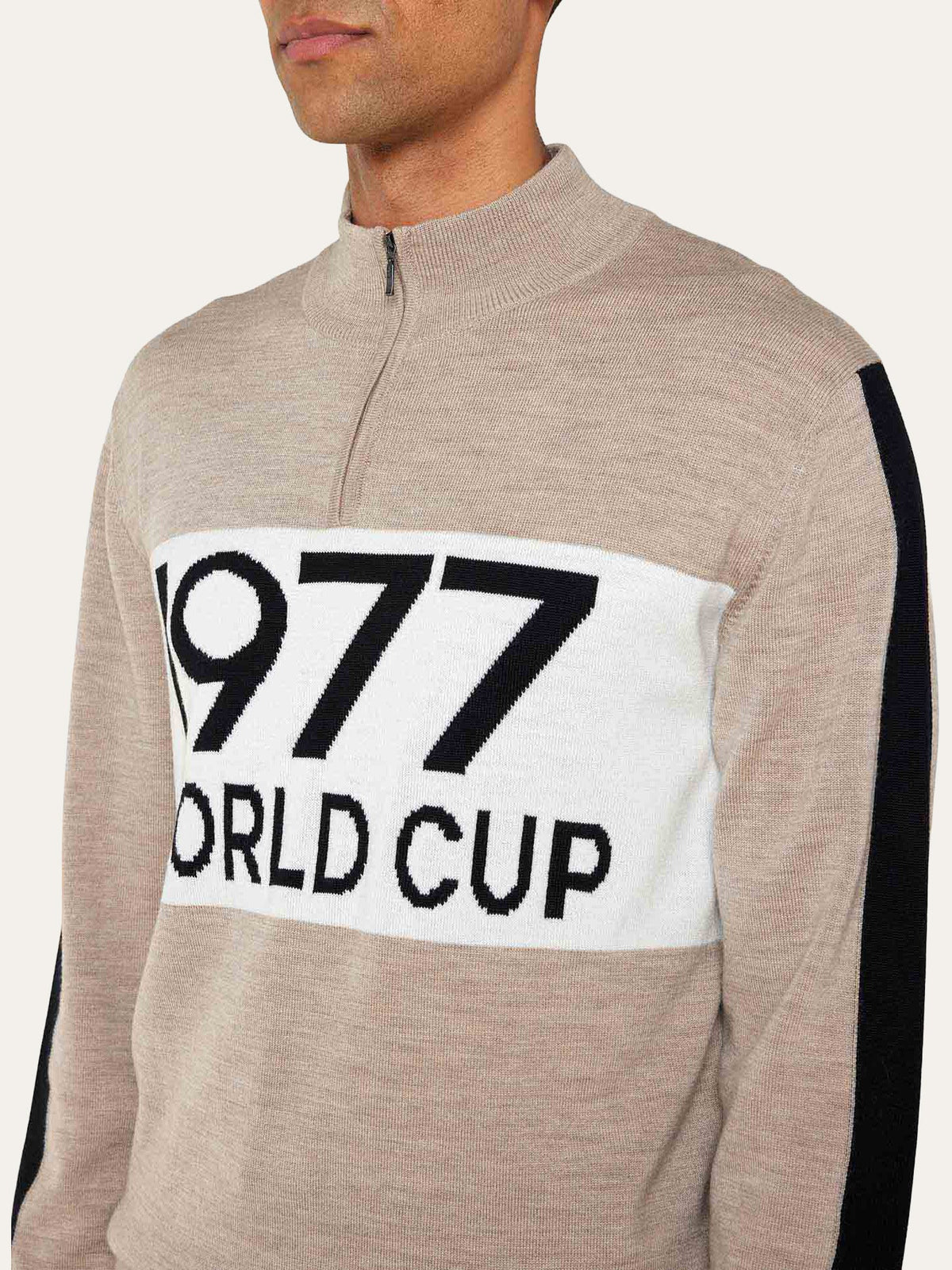 A person wearing the We Norwegians World Cup Zipup Men—a beige half-zip sweater with bold black and white “1977 WORLD CUP” lettering across the chest, inspired by 70s skiing style and classic merino wool ski sweaters.