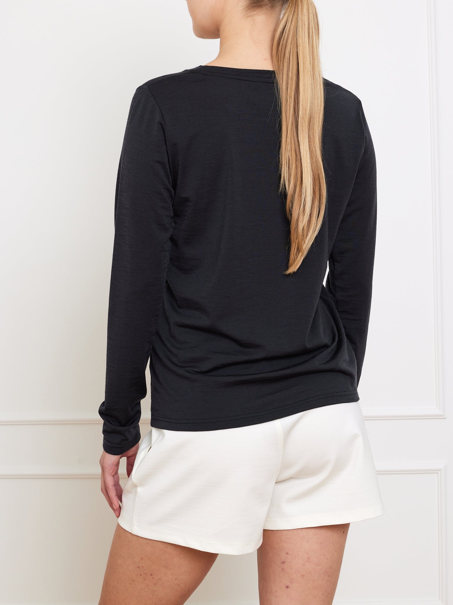 With long blonde hair, a person stands facing away in front of a white wall, wearing We Norwegians' Berg LongSleeve Women shirt in sleek black paired with white shorts.