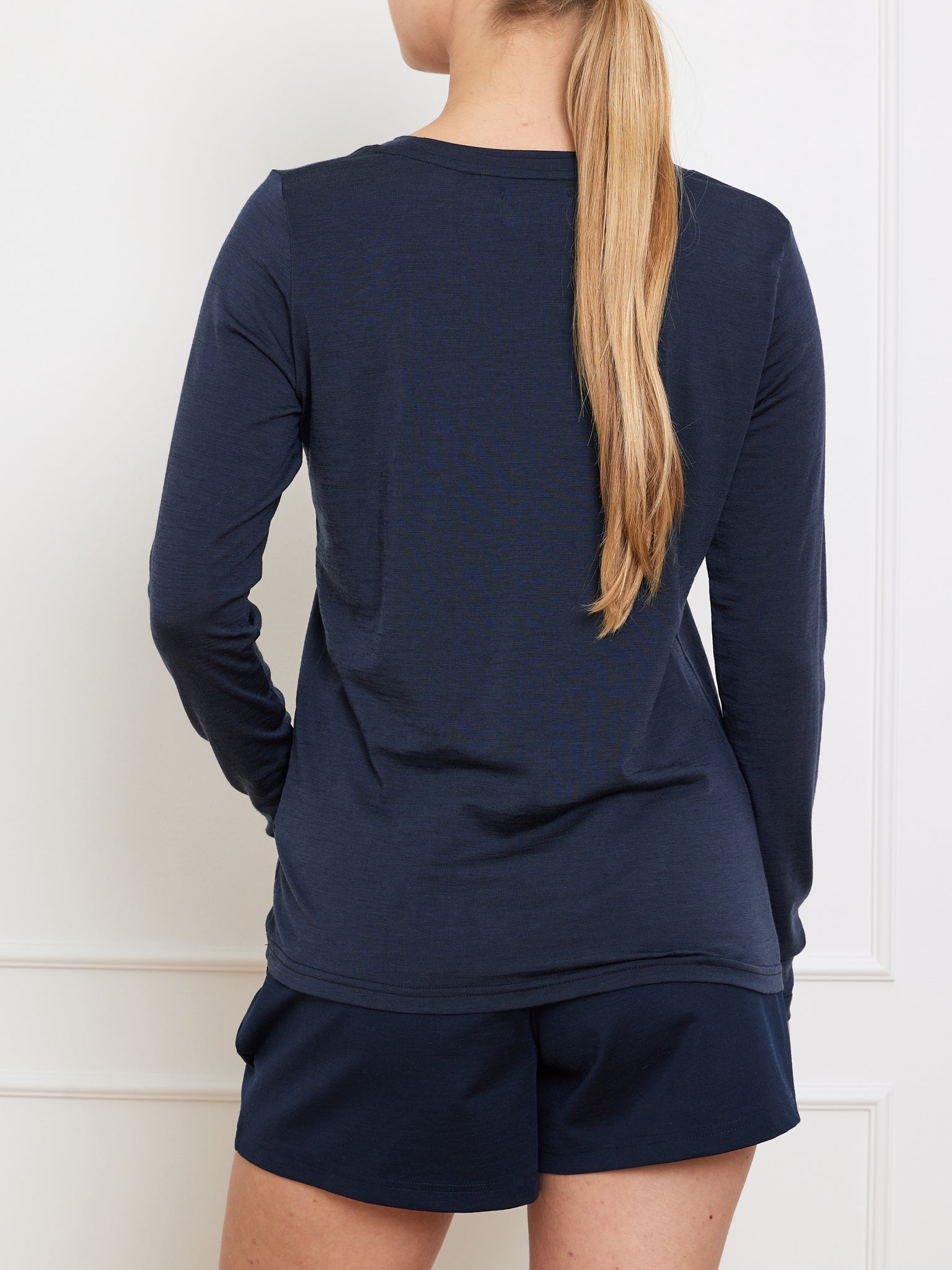 A person with long blonde hair is facing away from the camera, showcasing the We Norwegians' Berg LongSleeve Women shirt in navy blue, paired with matching shorts against a white background.